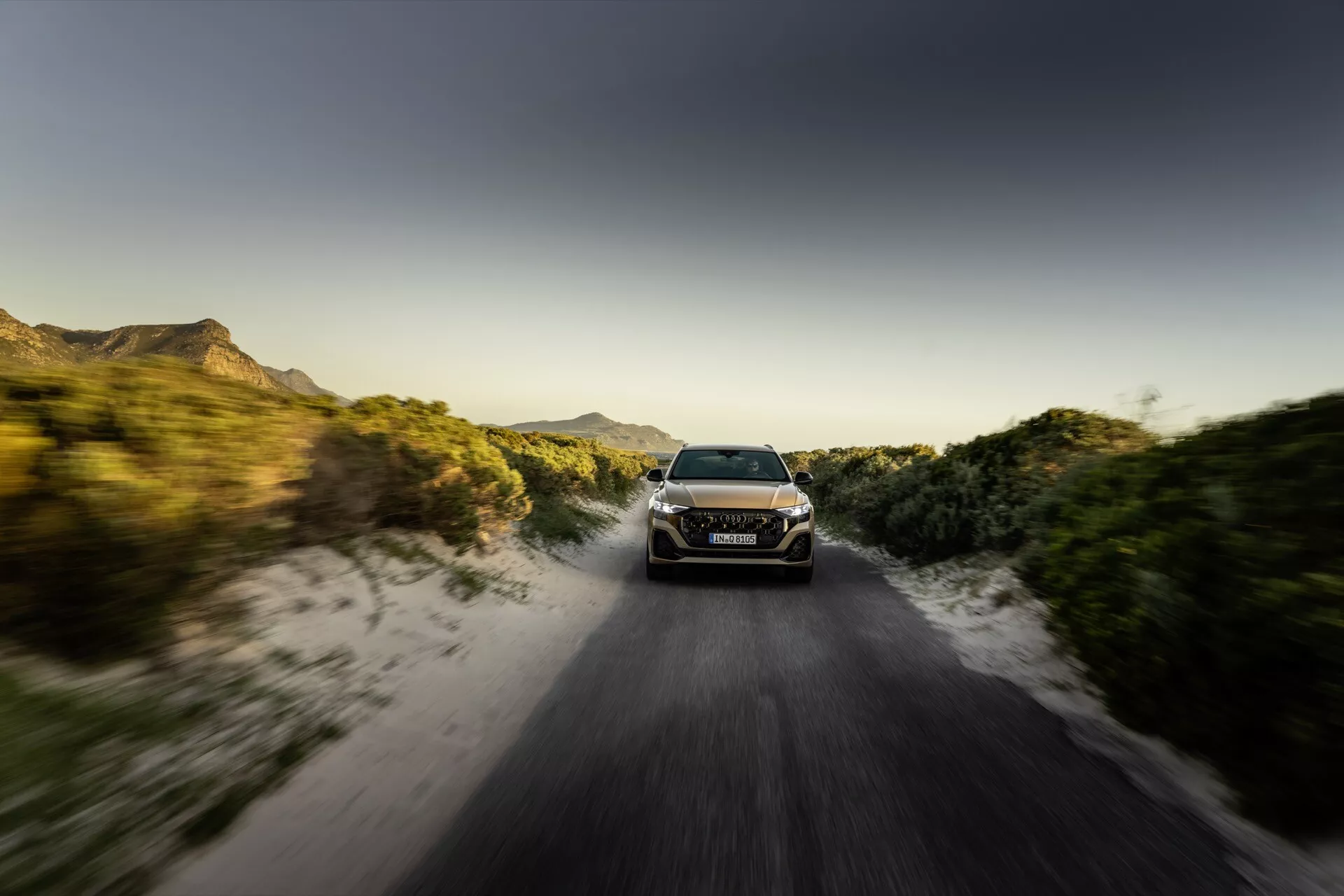 Audi Q8 2023 (4M-Facelift) w dynamicznym ruchu na nieutwardzonej drodze o wschodzie lub zachodzie słońca z efektem zamazania ruchu, prezentujące jego reflektory LED, elegancki aerodynamiczny design i zdolności terenowe wśród wiejskiego otoczenia