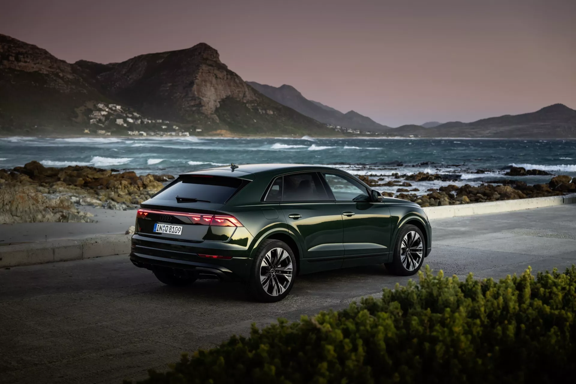 Audi Q8 2023 (4M-Facelift) zaparkowane w malowniczym nadmorskim miejscu o świcie lub zmierzchu, prezentujące widok z tylnej kwarty, ciemnozielone metaliczne wykończenie i wieloramienne felgi aluminiowe na tle spokojnego krajobrazu górskiego