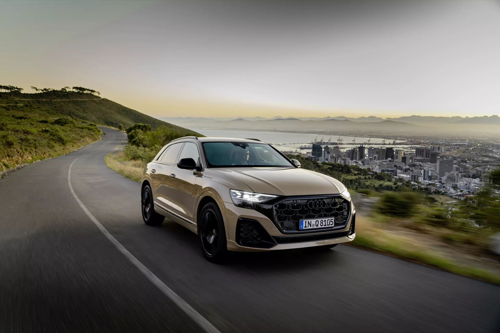 Luksusowy SUV crossover Audi Q8 2023 (4M-Facelift) w ruchu na krętej drodze, prezentujący sportowy design, reflektory LED, duże felgi aluminiowe i panoramiczny dach, na tle miasta i gór w miękkim świetle porannym lub popołudniowym.