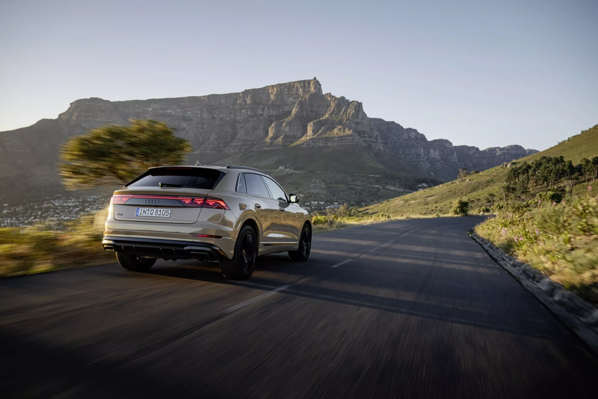 Model Audi Q8 4M facelift 2023 w dynamicznym ruchu na krętej drodze, podkreślający jego nowoczesny design SUV i zaawansowane funkcje z perspektywy trzech czwartych z tyłu, z malowniczym górskim tłem o zachodzie słońca