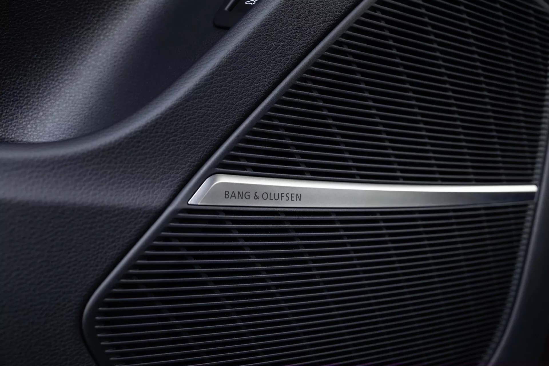 Zbliżenie na kratkę głośnika Bang & Olufsen we wnętrzu Audi Q8 2023 (4M-Facelift), przedstawiające drobny wzór siatki wysokiej jakości systemu audio i polerowane metaliczne logo na czarnym tle.