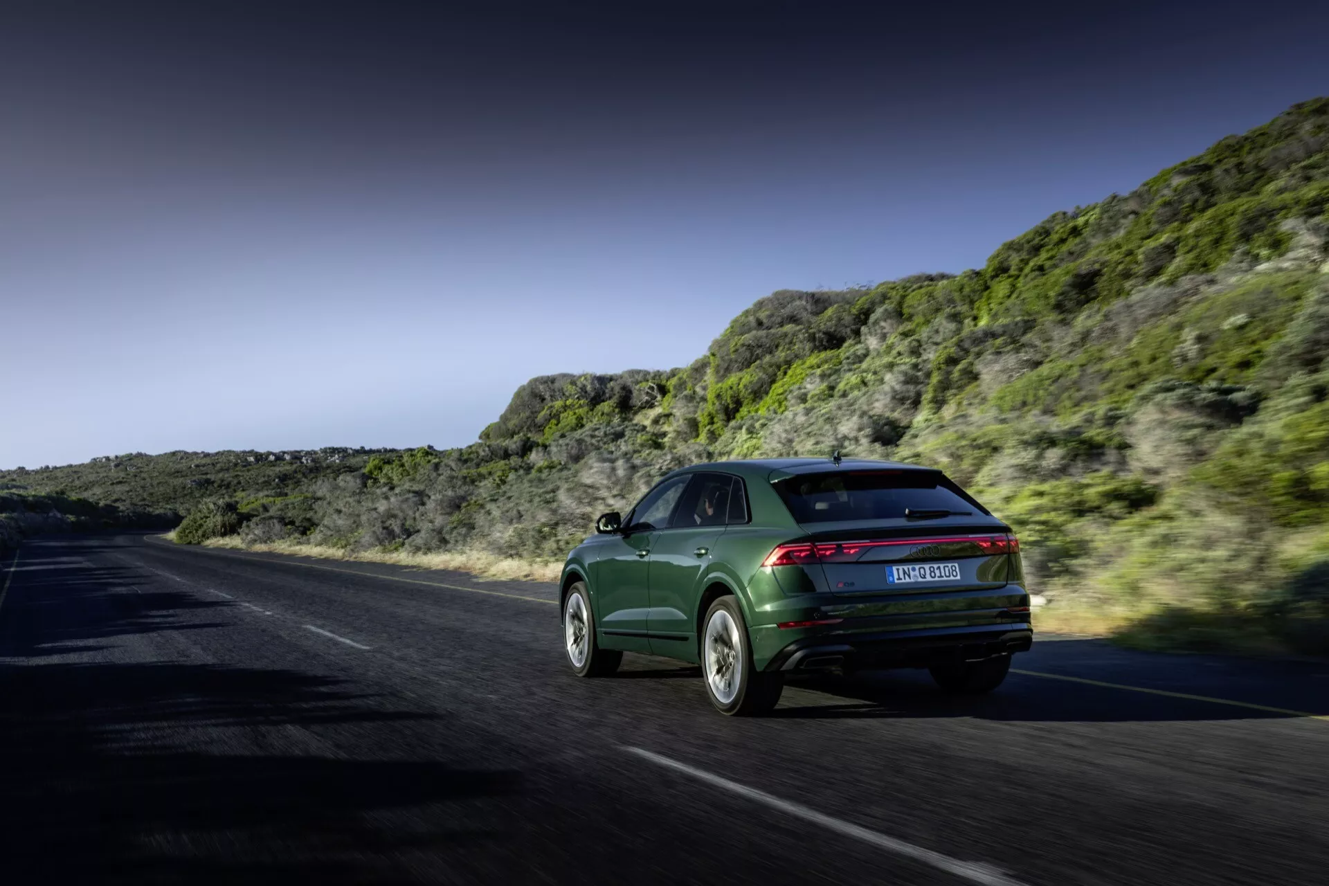 Audi Q8 2023 (4M-Facelift) w dynamicznym ruchu na przybrzeżnej lub górskiej drodze, prezentujący swój styl sportback, eleganckie tylne światła i wyrafinowane koła ze stopów lekkich na tle dramatycznego krajobrazu