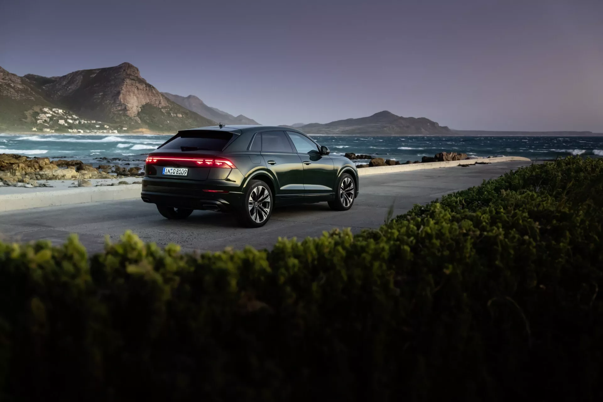 Audi Q8 4M-Facelift 2023 w metalicznym wykończeniu, uchwycone o świcie lub zmierzchu w naturalnym nadmorskim otoczeniu, podkreślające widok z tyłu z trzech kwadransów z tylnymi lampami LED, podwójnym wydechem i felgami ze stopu o wielu ramionach.