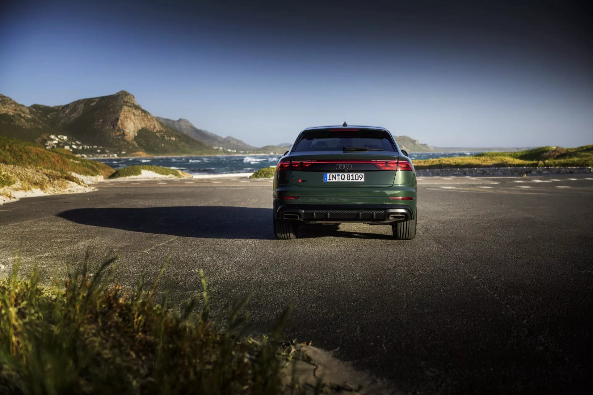 Tylny widok modelu Audi Q8 4M Facelift 2023, prezentujący tylne światła LED, akcenty chromowane i podwójny wydech na malowniczej drodze przybrzeżnej