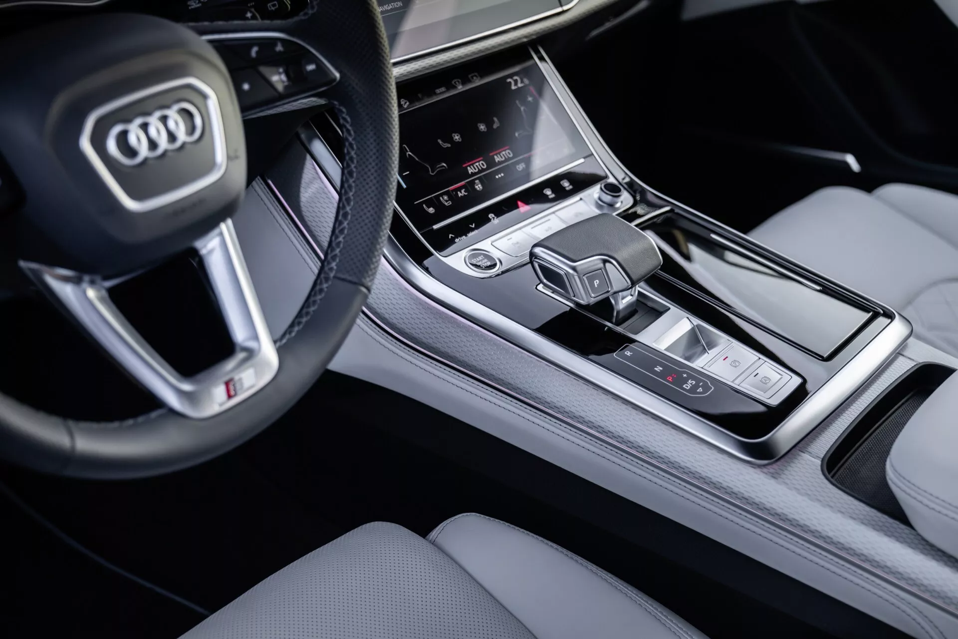 Zbliżony widok na konsolę środkową i część kierownicy Audi Q8 2023 (4M-Facelift) z perspektywy pasażera, podkreślający przyciski na wielofunkcyjnej kierownicy, centralny ekran dotykowy systemu infotainment, obszar kontroli klimatyzacji i nowoczesną dźwignię zmiany biegów o wysokim standardzie wykończenia wnętrza.