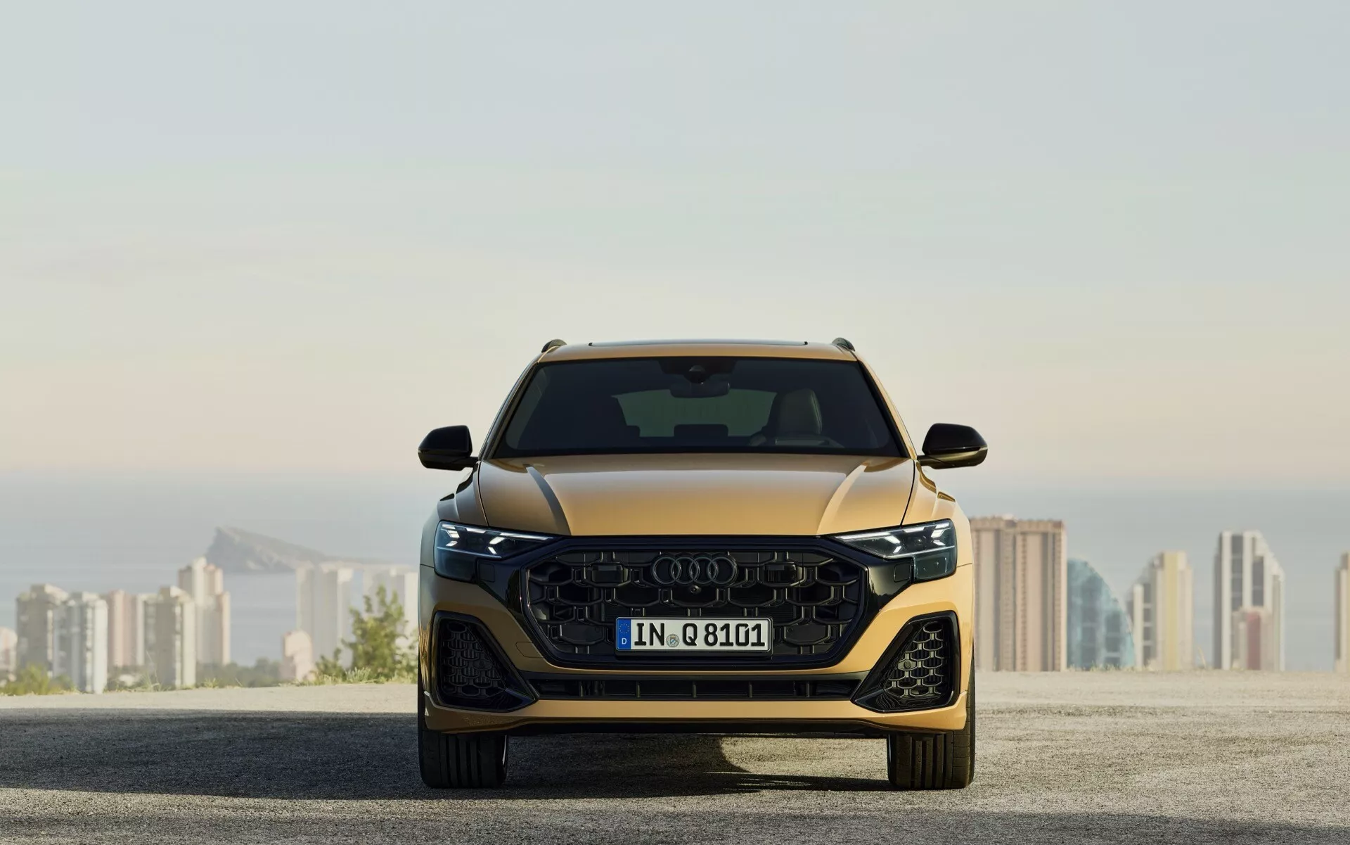 Pełny frontowy widok Audi Q8 (4M-Facelift) 2023 ukazujący jego aerodynamiczny design, dużą atrapę z logo z czterema pierścieniami, smukłe jednostki reflektorów LED i zintegrowane zaawansowane systemy wspomagania kierowcy, na tle rozmazanego krajobrazu miejskiego