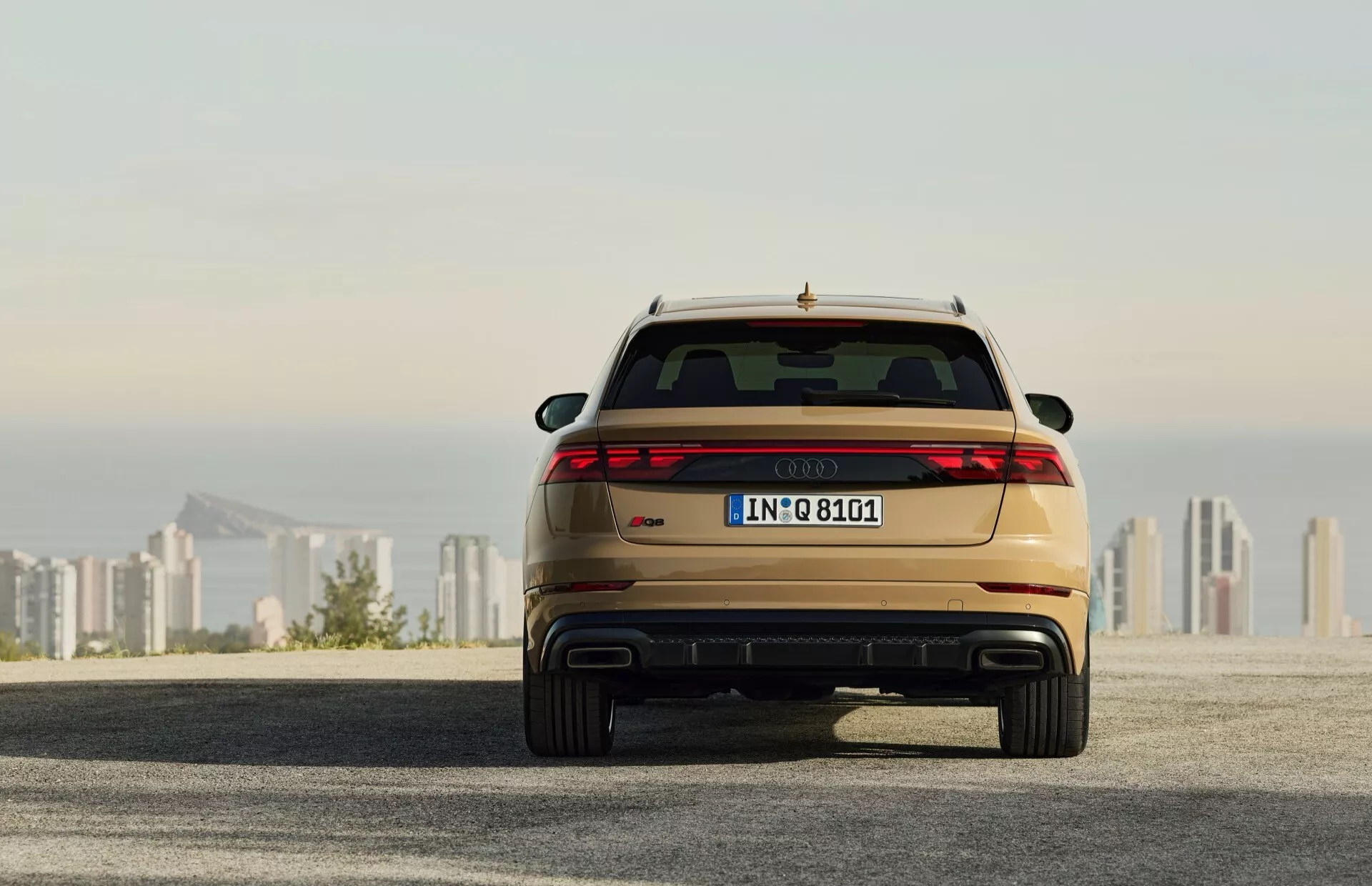 Tył Audi Q8 2023 (4M-Facelift) pokazujący tylne światła LED, podwójny wydech i metaliczny lakier, zaparkowany na asfalcie z widokiem na linię horyzontu miasta w tle o wschodzie/zachodzie słońca