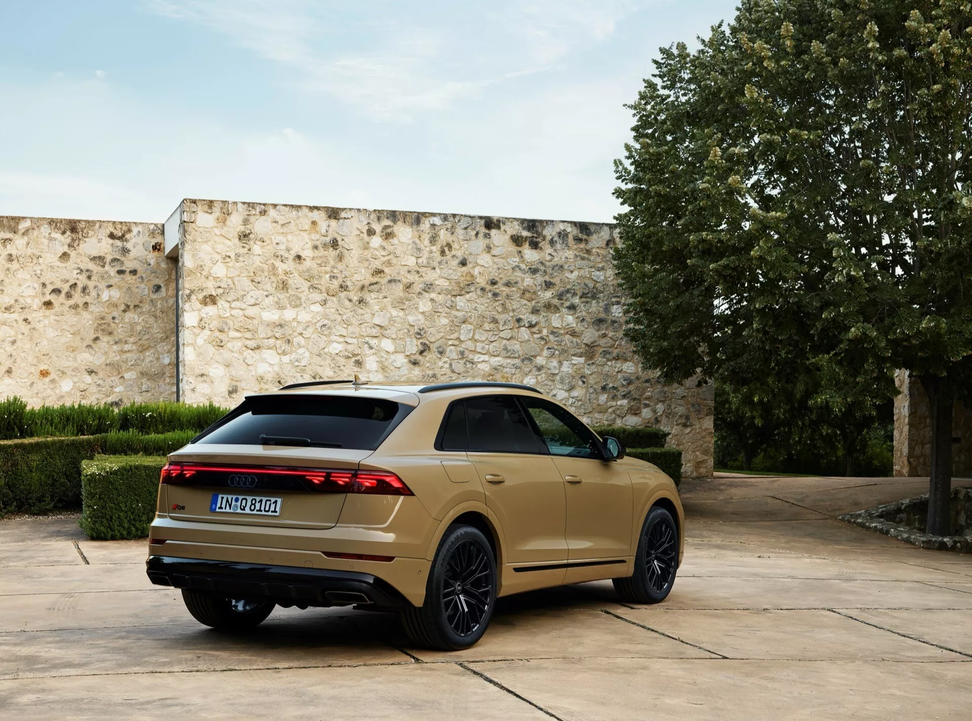 Model Audi Q8 4M po liftingu z 2023 roku zaparkowany na zewnątrz, uchwycony z tylnego kąta trzech czwartych, podkreślający jego tylne światła LED, ciemne felgi aluminiowe z wieloma szprychami, opadający dach i podwójne rury wydechowe na tle kamiennego dziedzińca