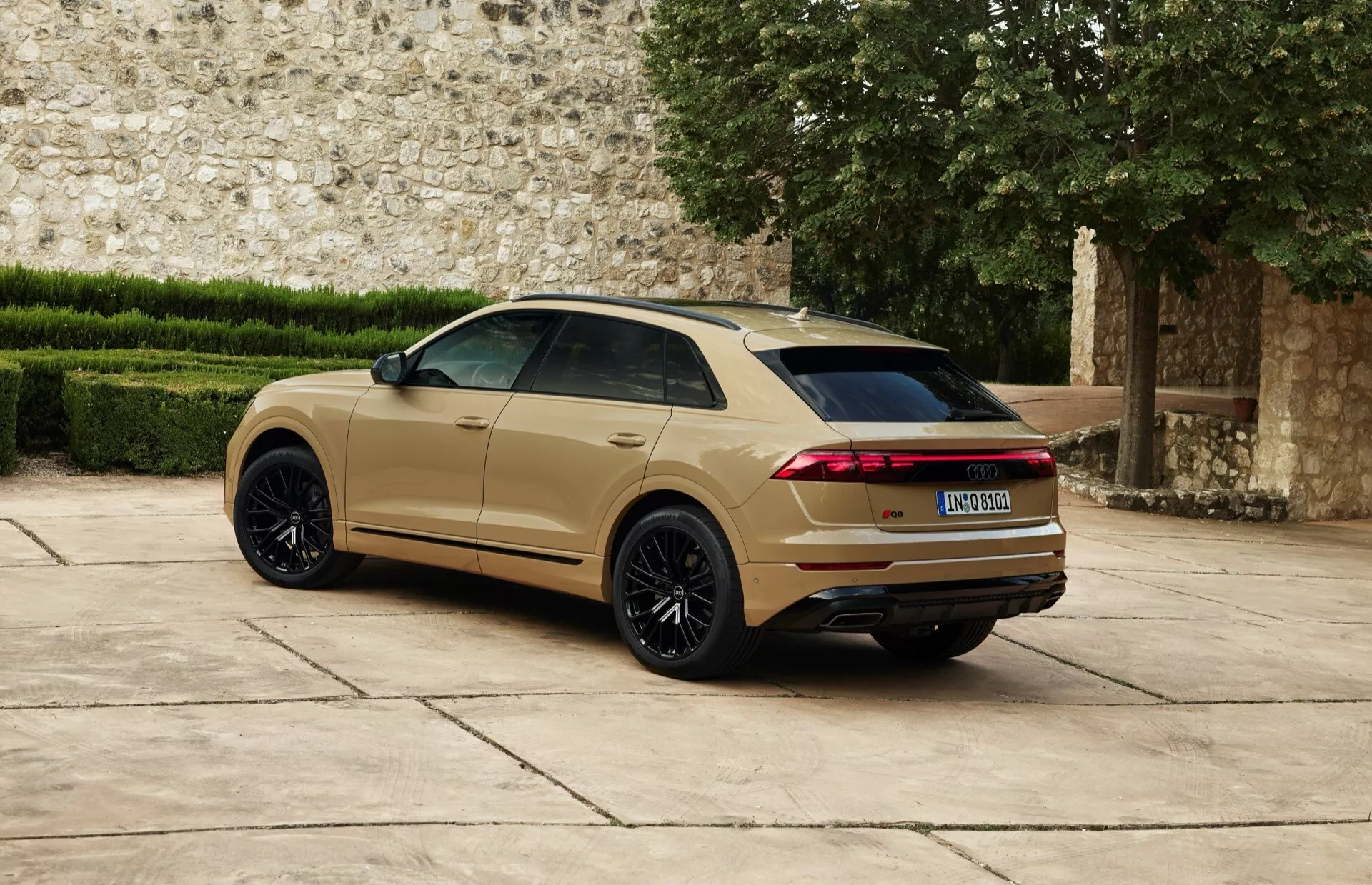 Audi Q8 2023 (4M-Facelift) pokazane z tyłu z perspektywy trzech czwartych z dużymi czarnymi kołami ze stopu wieloramienne, metalicznym złoto-beżowym lakierem, wydłużonymi poziomymi tylnymi światłami LED, tylnym spojlerem i podwójnym systemem wydechowym na tle historycznego muru kamiennego