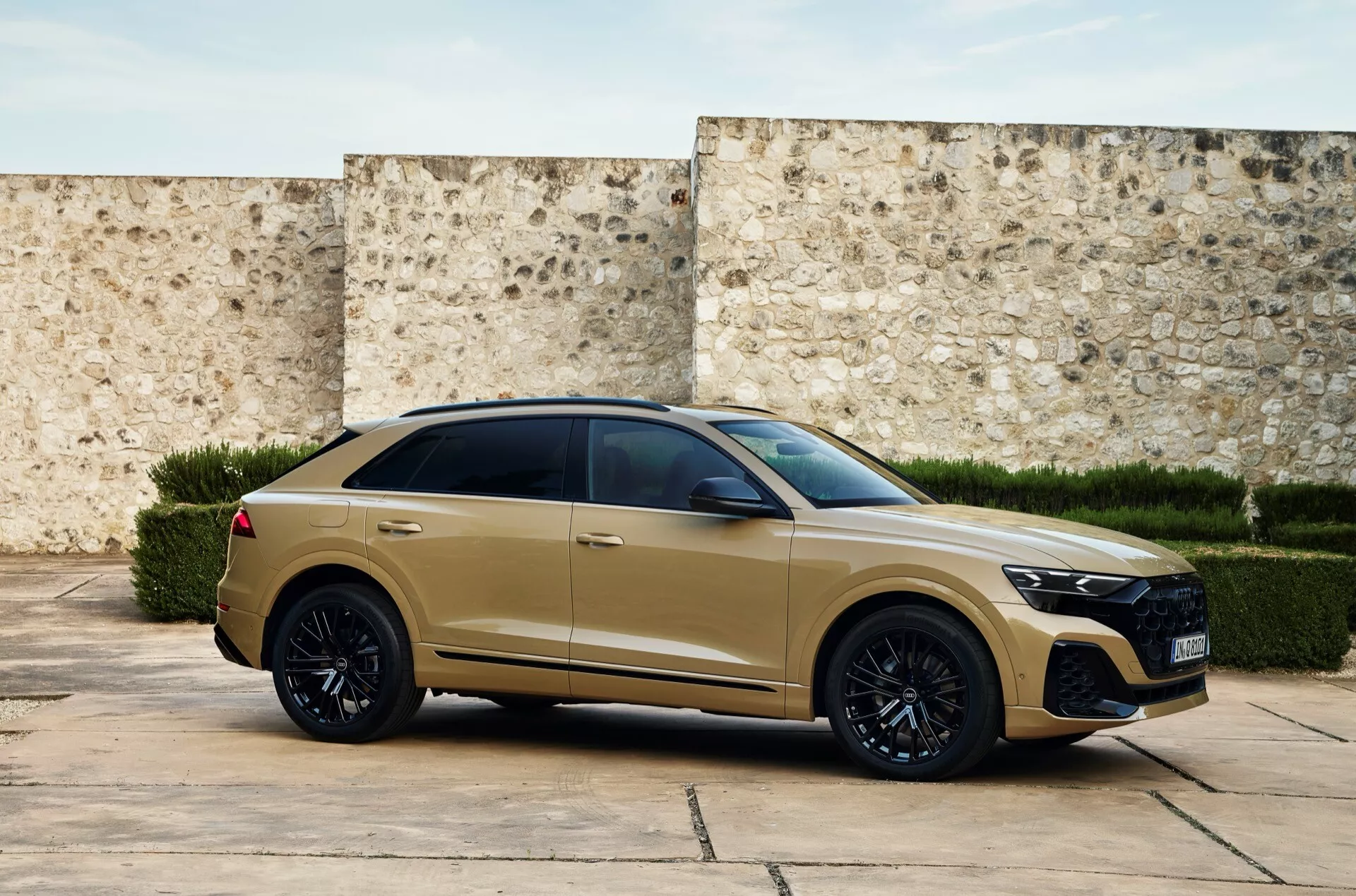 Widok z przodu pod kątem trzech czwartych nowoczesnego Audi Q8 2023 (4M-Facelift) w matowym złotym lub beżowym wykończeniu, z dużym grillem w wzór plaster miodu, reflektorami LED, sportowym zderzakiem przednim, czarnymi kołami ze stopów, oponami o niskim profilu, chowanymi klamkami, przyciemnianymi szybami, linia dachu na kształt coupé, muskularna linia ramion z kątowymi światłami tylnymi i spojlerem dachowym, na tle teksturyzowanej ściany sugerującej historyczne lub współczesne otoczenie.