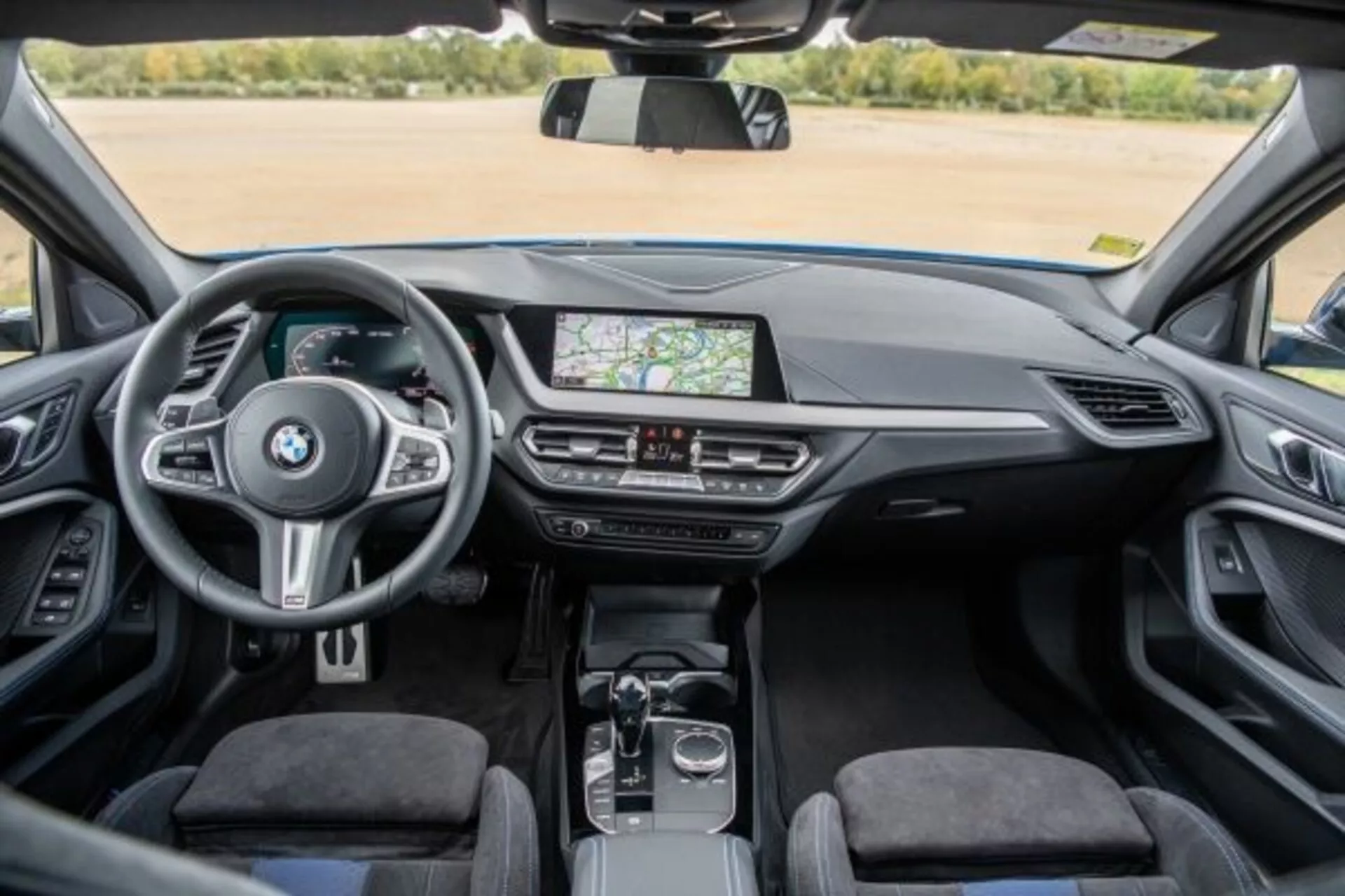Wnętrze BMW serii 1 M Performance 2019 z perspektywy tylnej kanapy ukazujące deski rozdzielczej, konsoli środkowej i kierownicy z zamontowanymi przyciskami, system infotainment z ekranem dotykowym wyświetlającym nawigację GPS, klimatyzację, automatyczną dźwignię zmiany biegów, pokrętło i touchpad na konsoli środkowej, tapicerkę premium oraz jasne i dobrze oświetlone wnętrze