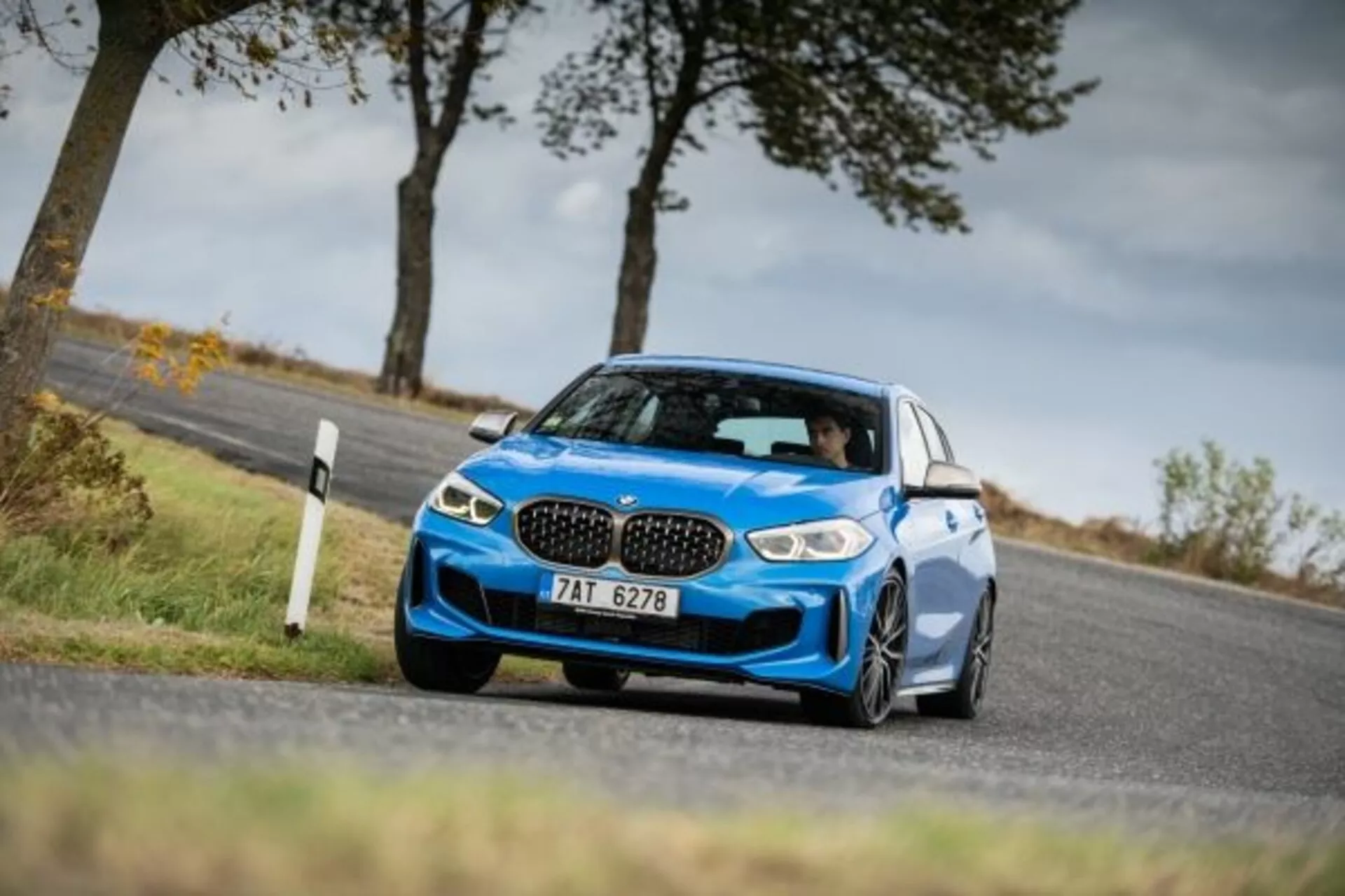 Niebieski sportowy sedan BMW Serii 1 M Performance z 2019 roku uchwycony z niskiej i skośnej perspektywy boczno-frontalnej podkreślającej jego agresywną przednią fasadę z ikonicznymi grillami nerkowymi, eleganckimi reflektorami LED, dużymi wlotami powietrza do chłodzenia osiągów, niskim zawieszeniem sportowym, w ruchu wokół zakrętu ukazującym dynamiczną obsługę, z aerodynamicznymi lusterkami bocznymi, dużymi kołami ze stopów i oponami o niskim profilu podkreślającymi sportowy status i zdolności przyczepności.
