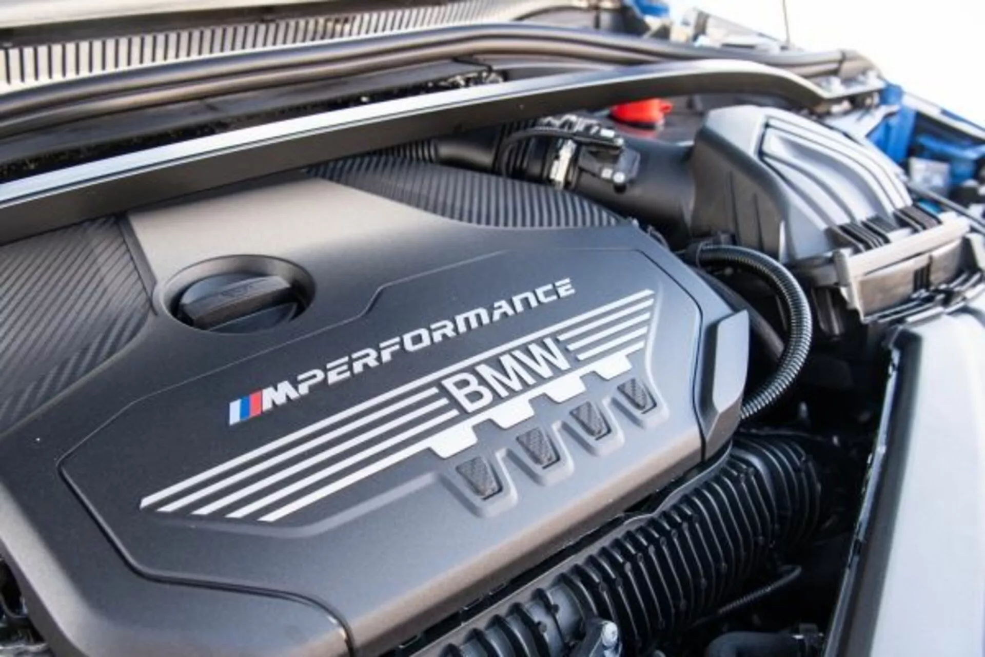 Zbliżenie na komorę silnika BMW Serii 1 M Performance z 2019 roku z pokrywą silnika ozdobioną logo M w charakterystycznych kolorach niebieskim, purpurowym i czerwonym, podkreślająca sportowy, wysokowydajny projekt.