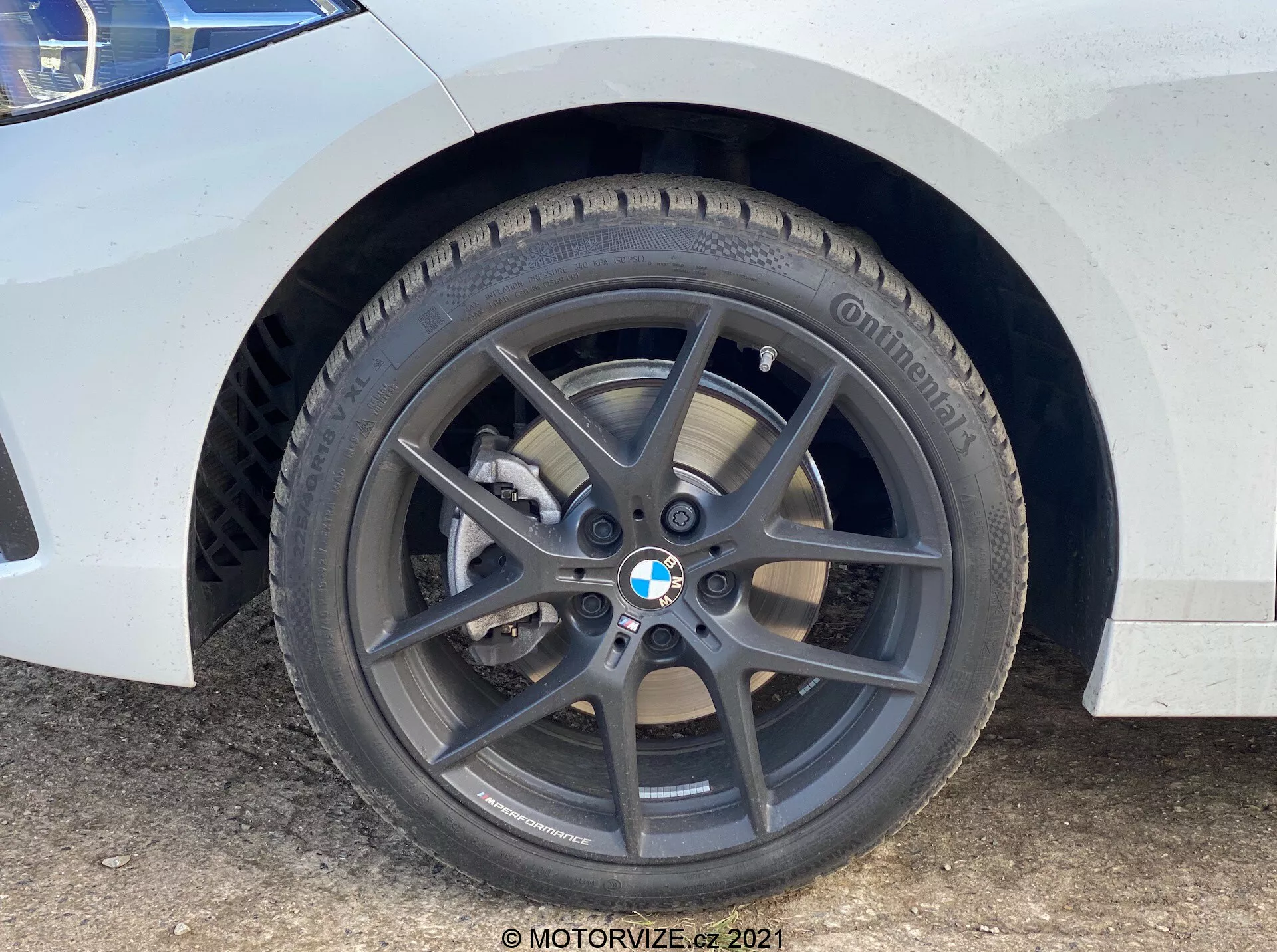 Zbliżenie na przednie koło i błotnik BMW serii 1 2019 (BMW-116i). Ujęcie z perspektywy ziemi podkreśla ciemnoszarą, pięcioramienną felgę aluminiową z logo BMW, markową oponę Continental z widocznym bieżnikiem, wentylowany tarczowy układ hamulcowy oraz srebrny jednopiścienowy zacisk hamulca. Biała karoseria z brudem i plamami wody wskazuje na stan użycia.