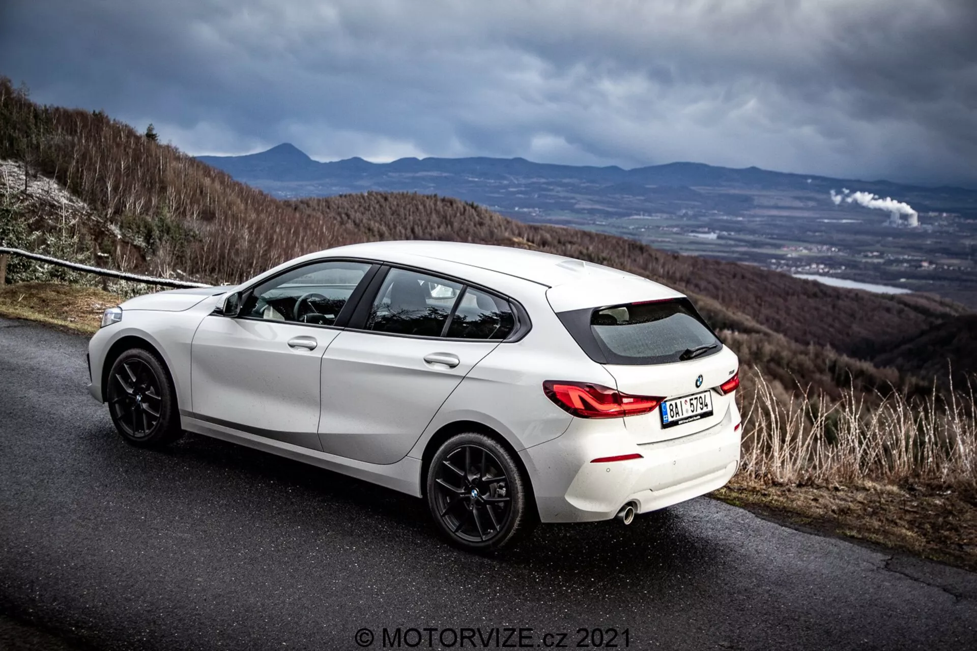 Skośny widok tylno-boczny białego hatchbacka BMW 116i z 2019 roku na asfaltowej drodze z dramatycznym pochmurnym niebem. Obraz podkreśla tylną stylistykę pojazdu, tylne nadkola, ciemne felgi aluminiowe, podwójny układ wydechowy i profil boczny, w tym tylnych i przednich drzwi kierowcy. Otoczenie sugeruje drogę na wzgórzu lub górze, podkreślając sportowe i dynamiczne cechy samochodu. Obecny znak wodny fotografa 'MOTORWIZE.cz 2021'.