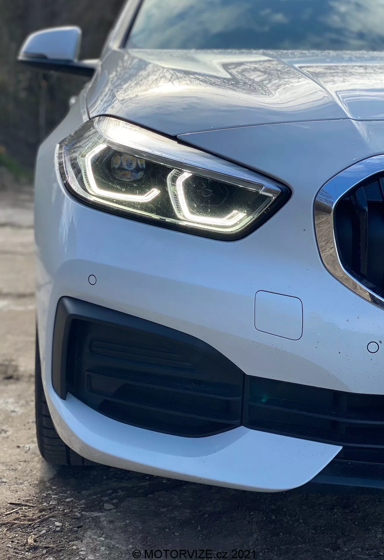 Zbliżenie przedniej lewej części samochodu BMW Serii 1 z 2019 roku (BMW 116i) podkreślające nowoczesny reflektor z charakterystycznymi światłami do jazdy dziennej LED, kratkę chłodnicy o wzorze plastra miodu z dużym wlotem powietrza, wyrafinowany aerodynamiczny design przedniego zderzaka i czujnik parkowania wbudowany w zderzak, prezentujące metaliczne wykończenie premium i odblaskowe nadwozie.