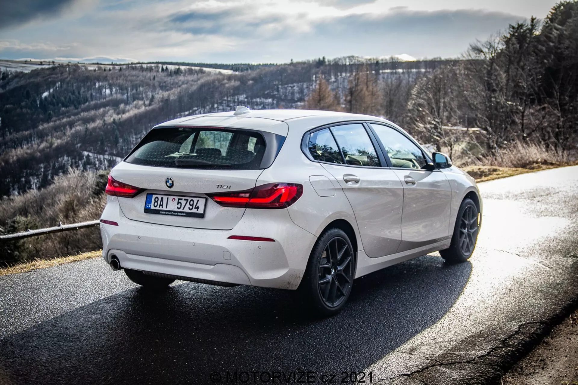 Dynamiczny widok tylno-trzy-czwarte białego BMW 116i 2019, pokazujący prawą stronę i tył pojazdu w świetle dziennym z wyraźnymi promieniami słońca i cieniami. Obraz podkreśla detale projektu, w tym czarne akcenty na tylnym dyfuzorze, felgach i obudowach lusterek bocznych, uzupełnione o aluminiowe felgi i podwójny układ wydechowy, sugerując sportowszą wersję. Droga zakręca w prawo, z górzystym lub pagórkowatym tłem, idealna dla entuzjastów jazdy.
