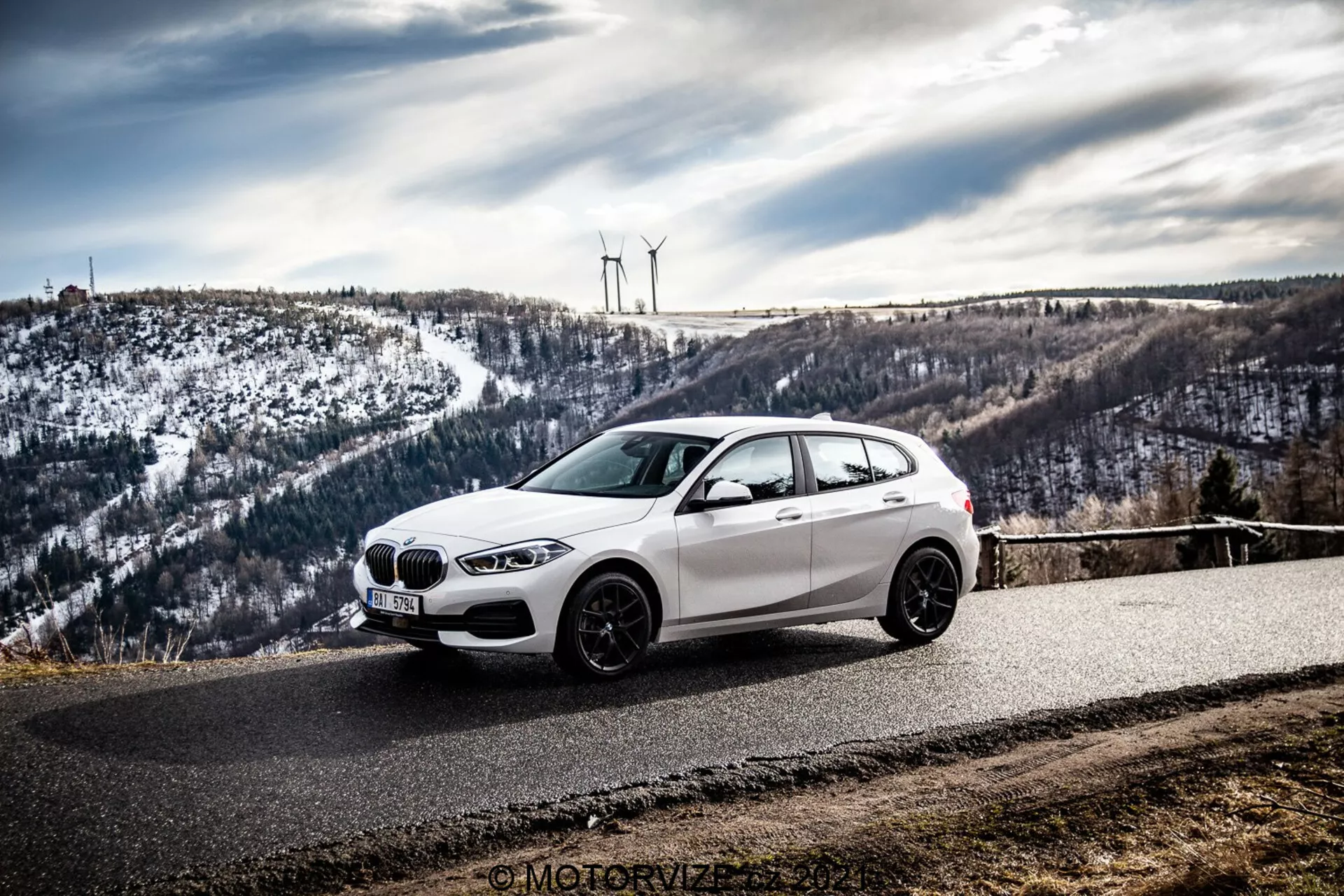 Biały hatchback BMW, prawdopodobnie BMW 1er-Reihe 2019 (BMW 116i), stoi na zakrzywionej asfaltowej drodze otoczonej pagórkowatym, śnieżnym krajobrazem z turbinami wiatrowymi. Zdjęcie zostało wykonane pod kątem boczno-przednim, podkreślając przedni grill, reflektory, linie aerodynamiczne i felgi aluminiowe auta na tle zachmurzonego nieba.