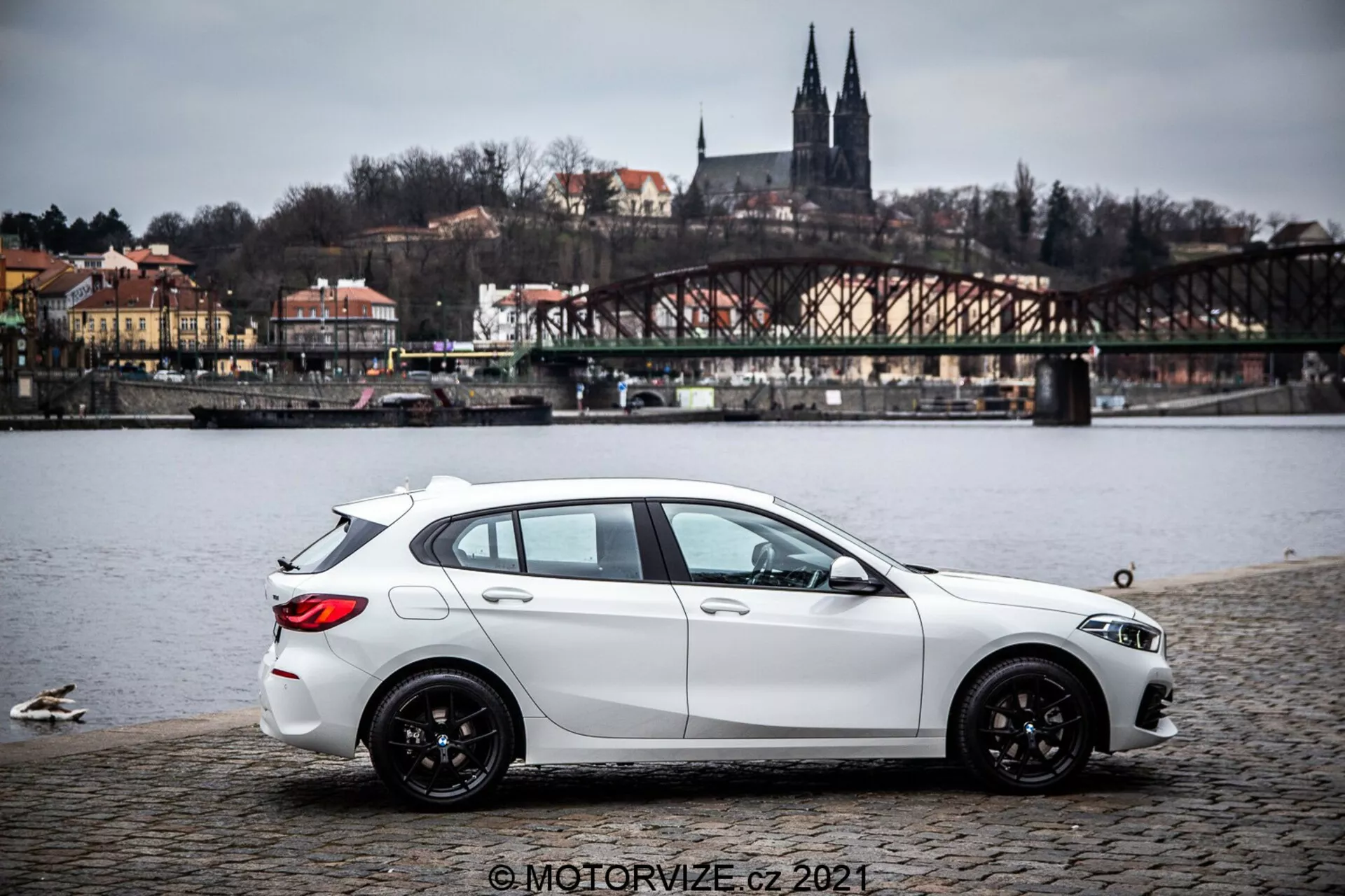 Biały hatchback BMW Serii 1 z 2019 roku (BMW-116i) zaparkowany na powierzchni z kostki brukowej w pobliżu zbiornika wodnego z kaczkami, ukazujący profil boczny z lekkim widokiem tylnego ćwiartki. Samochód ma dynamiczne rzeźbione linie, opadającą linię dachu, czarne wieloramienne felgi aluminiowe oraz widoczne reflektory i lusterka boczne. W tle znajduje się most, budynki na wzgórzu i elementy architektury sugerujące europejskie tło. Oświetlenie jest rozproszone pod zachmurzonym niebem, a obraz zawiera znak wodny © MOTORVIZE.cz 2021.