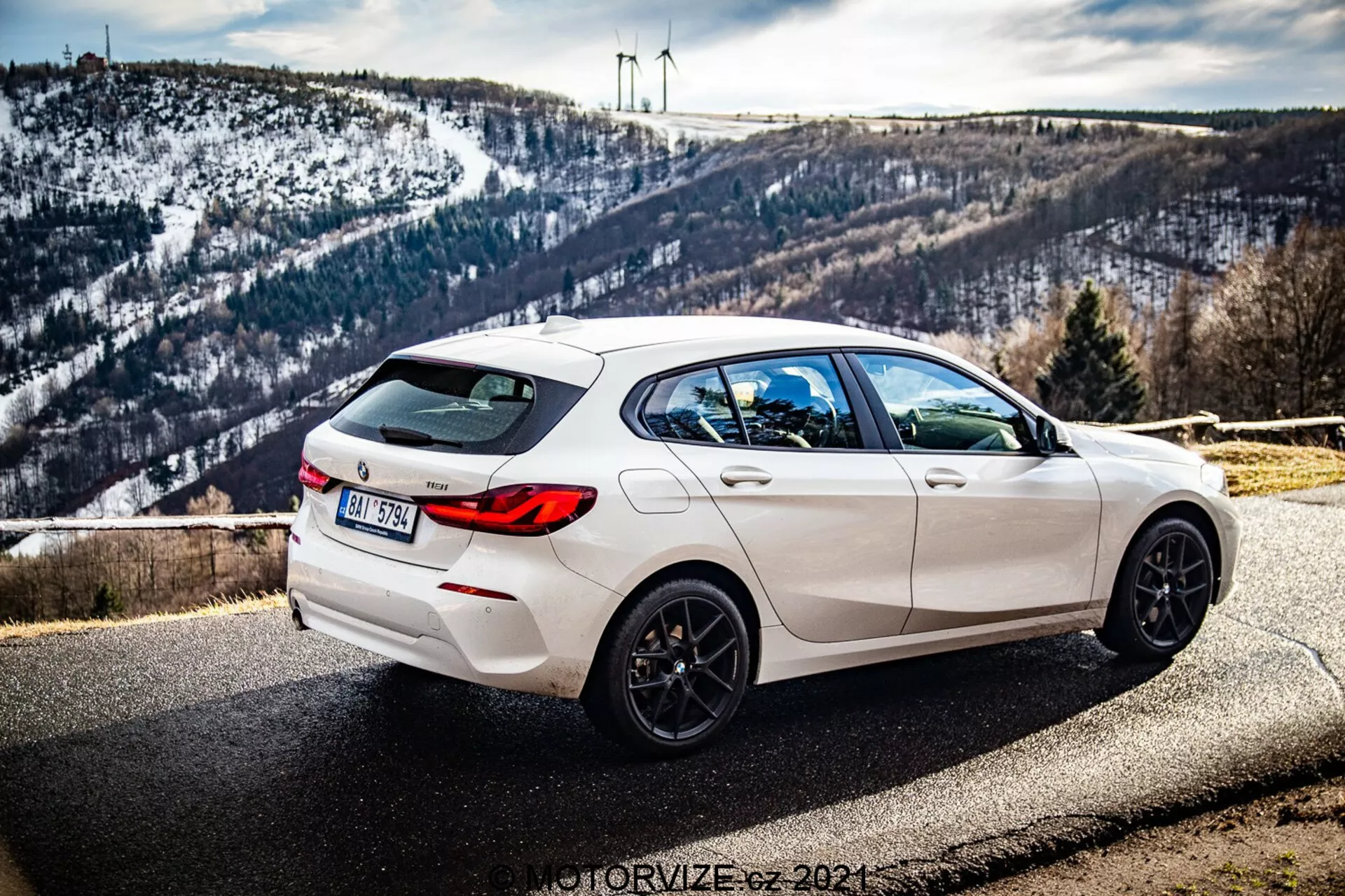 Obraz przedstawia białe BMW Serii 1 (BMW-116i) z 2019 roku zaparkowane na poboczu drogi z malowniczym tłem, na którym widać pagórkowaty, ośnieżony krajobraz oraz turbiny wiatrowe na horyzoncie. Zdjęcie wykonano z potrójnego kąta tylnego, ukazując wyraźnie prawą stronę pojazdu. Widok podkreśla eleganckie linie projektowe, tylny spojler, czarne obręcze pięcioramienne, podwójne wydechy i zaciemnione szyby z tyłu. Auto ma również tylnią wycieraczkę oraz zintegrowaną antenę typu 