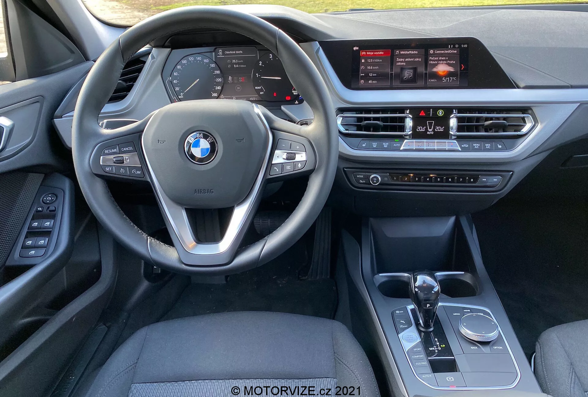 Widok wnętrza BMW Serii 1 (BMW 116i) 2019 z perspektywy fotela pasażera z przodu, ukazujący wielofunkcyjną kierownicę z zamontowanymi kontrolkami, cyfrowym zestawem wskaźników, dużym ekranem dotykowym systemu infotainment, systemem klimatyzacji, automatyczną dźwignią zmiany biegów, selektorem trybu jazdy i pokrętłem sterującym iDrive.