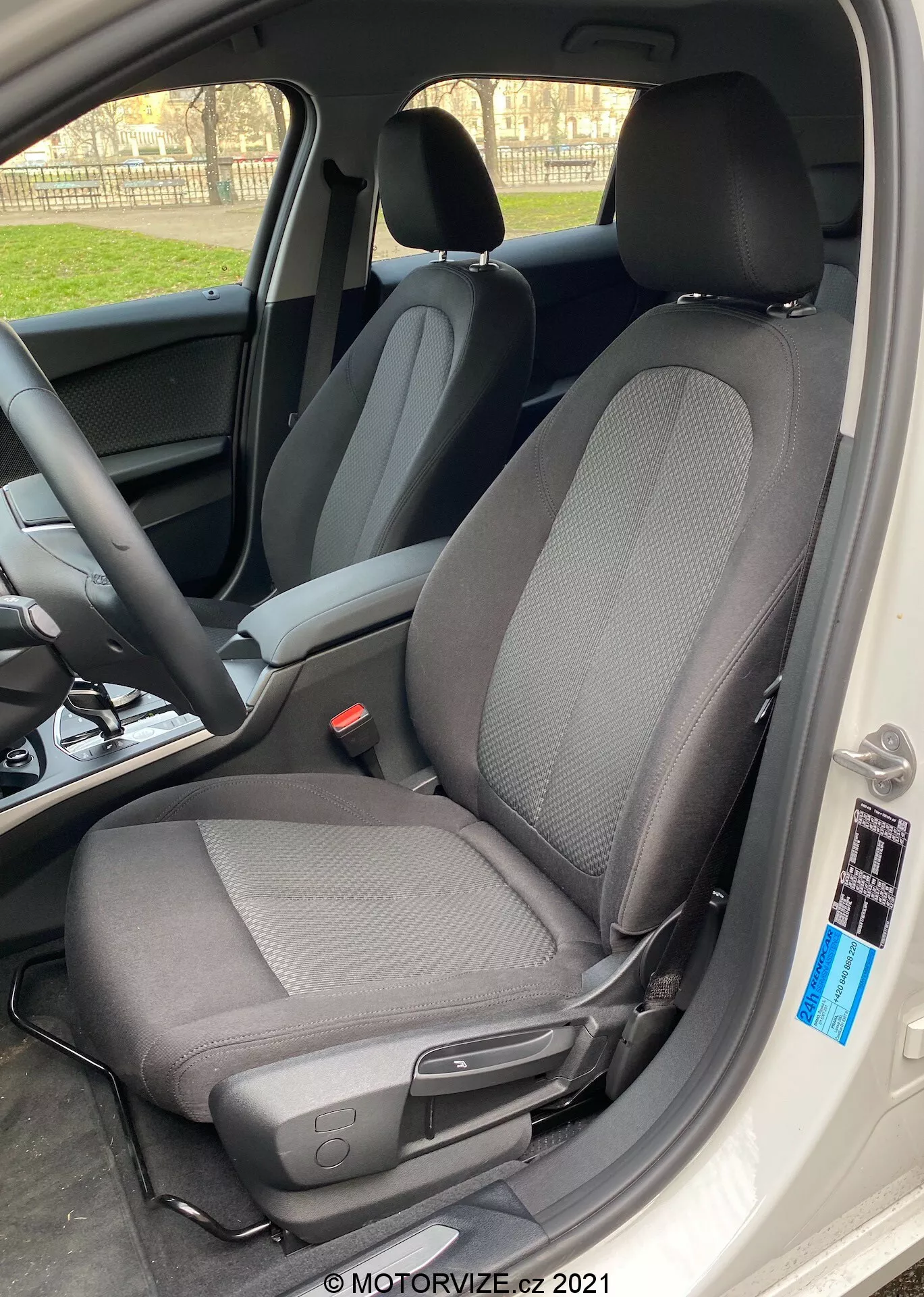 Zbliżenie wnętrza BMW 1er-Reihe 2019 (BMW-116i) koncentrujące się na fotelu kierowcy z nowoczesnym dwutonowym wzorem tkaniny, głównie czarnym z jaśniejszymi szarymi wzorami, wykonane pod kątem bocznym z szeroko otwartymi drzwiami samochodu. Fotel wyposażony jest w zagłówek ustawiony przeciwko światłu, tworząc efekt sylwetki wokół krawędzi, i zawiera dźwignię do ręcznego regulowania do przodu lub do tyłu, a także małe czarne pokrętło w pobliżu krawędzi wnętrza samochodu. Naturalne światło dzienne podkreśla ostre skupienie na fotelu, z rozmytym tłem ukazującym kierownicę i deskę rozdzielczą.