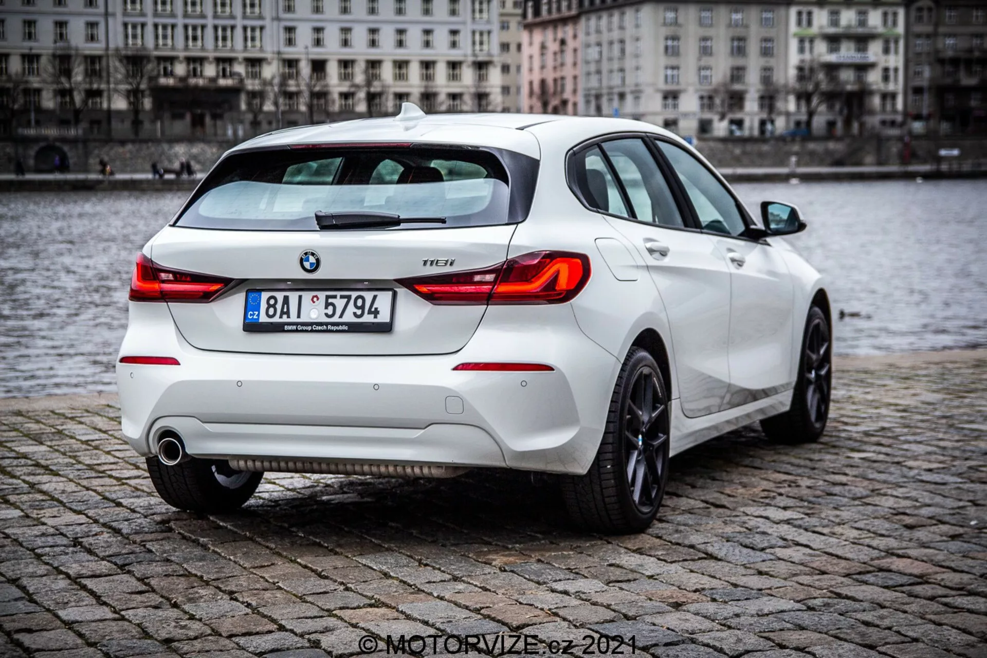 BMW Serii 1 z 2019 roku (116i) biały hatchback, przedstawiony z tylnego trójkątnego kąta z detalami, w tym tylne światła, pokrywa bagażnika, tylny zderzak, rury wydechowe i część tylnych kół, zaparkowany na mokrej nawierzchni z brukowcem blisko zbiornika wodnego. Zdjęcie podkreśla sportowy tylny spoiler, nowoczesne klastry świateł tylnych i widoczne emblematy BMW i poziom wyposażenia.