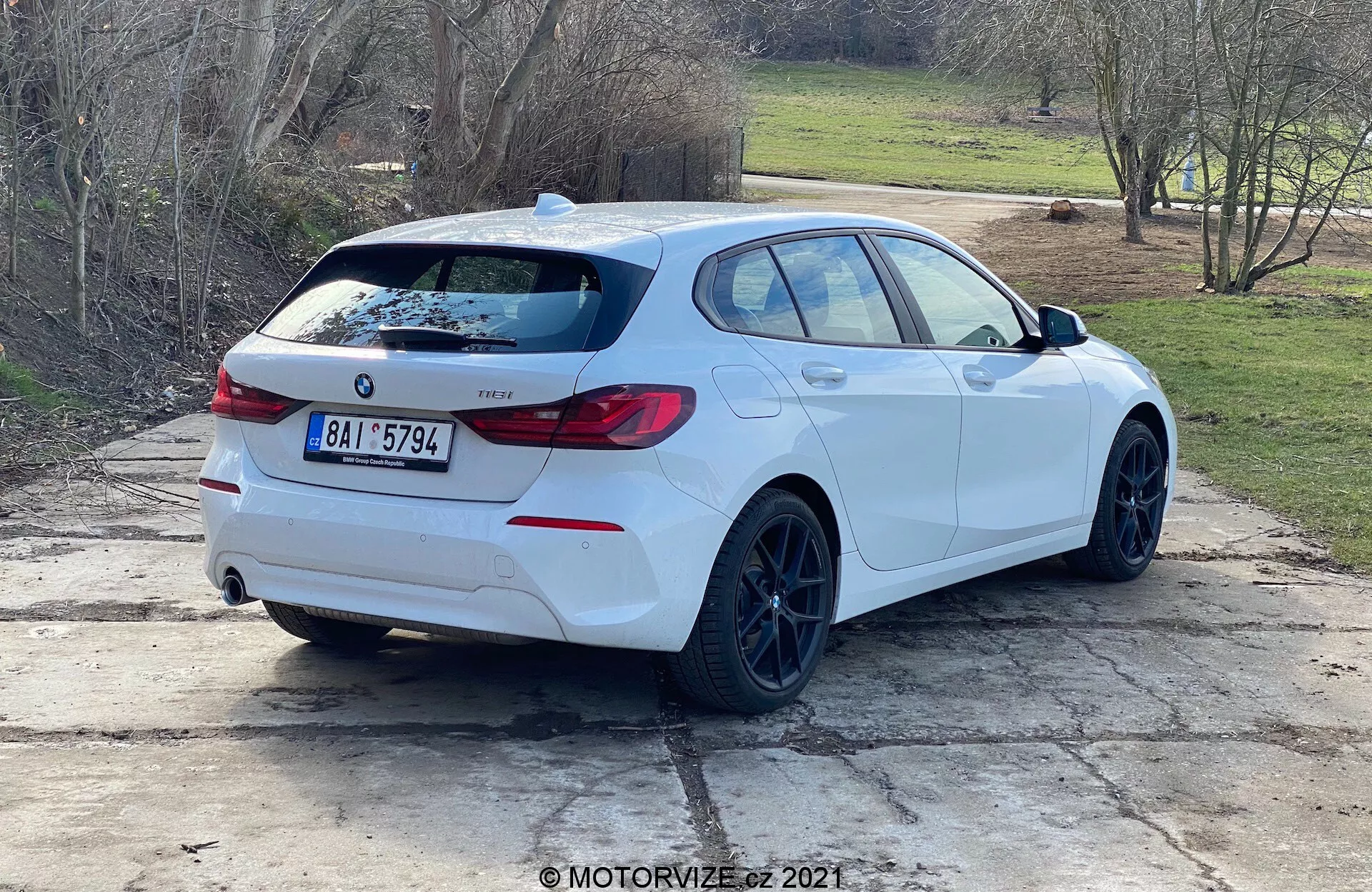 Widok z tyłu pod kątem na białe BMW serii 1 hatchback (model BMW 116i z 2019 roku) zaparkowane na zewnątrz w świetle dziennym. Profil boczny i tył pojazdu są wyraźnie widoczne, z emblematem BMW, oznaczeniem modelu, charakterystycznymi światłami tylnymi, tylnym spojlerem, czarnymi felgami, nadwoziem typu hatchback, podwójnym układem wydechowym, tylna wycieraczka, lekko przyciemnionymi szybami oraz lusterkami wstecznymi z zintegrowanymi kierunkowskazami. Samochód jest ustawiony na powierzchni betonowej z kępkami trawy i wiejskim lub podmiejskim tłem.