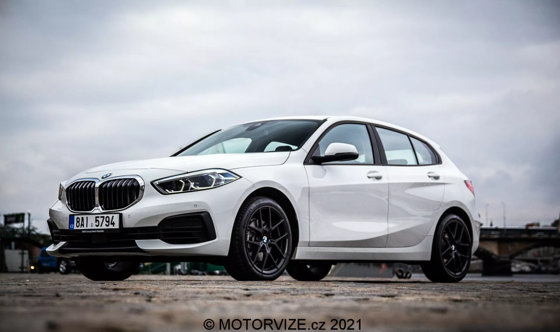 Boczny widok pod kątem na białe BMW Serii 1 2019 (BMW 116i), z wyraźnymi elementami designu, takimi jak krata w kształcie nerek, eleganckie reflektory LED i przedni zderzak z wlotami powietrza i możliwymi czujnikami radarowymi wspomagania kierowcy. Zdjęcie koncentruje się na felgach aluminiowych i dolnej części pojazdu, podkreślając sportowy, czarny wieloszprychowy design i wydajny hamulec tarczowy. Linie charakterystyczne i aerodynamiczne kształty hatchbacka dodatkowo podkreślają jego nowoczesny i dynamiczny wygląd zewnętrzny.