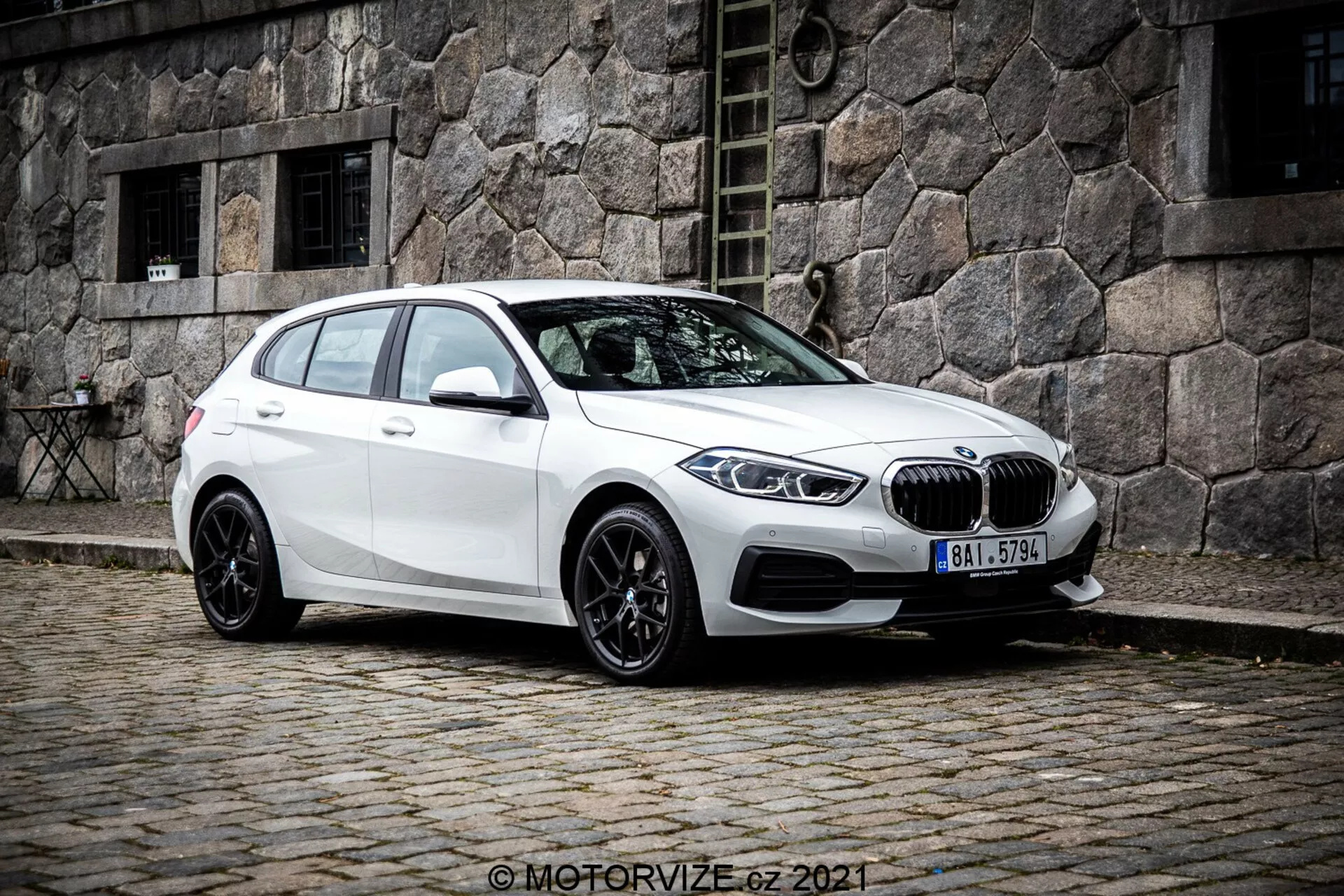 Białe BMW 1er-Reihe 2019 (BMW 116i) hatchback widziane pod kątem trzech czwartych z przodu, ukazujące lewą stronę pojazdu. Zdjęcie, zrobione z nieco niższego kąta, podkreśla sylwetkę i linie projektowe samochodu. Auto jest ustawione na brukowanej ulicy z zabytkowym kamiennym budynkiem w tle, tworząc klasyczną miejską scenę. Charakterystyczna krata nerkowa i emblemat BMW są wyraźnie widoczne z przodu, otoczone eleganckimi, kątowymi reflektorami LED. Samochód posiada wieloramienne czarne felgi aluminiowe, opadającą linię dachu, lusterka boczne w kolorze nadwozia z wbudowanymi kierunkowskazami oraz przyciemniane szyby dla tylnych pasażerów.