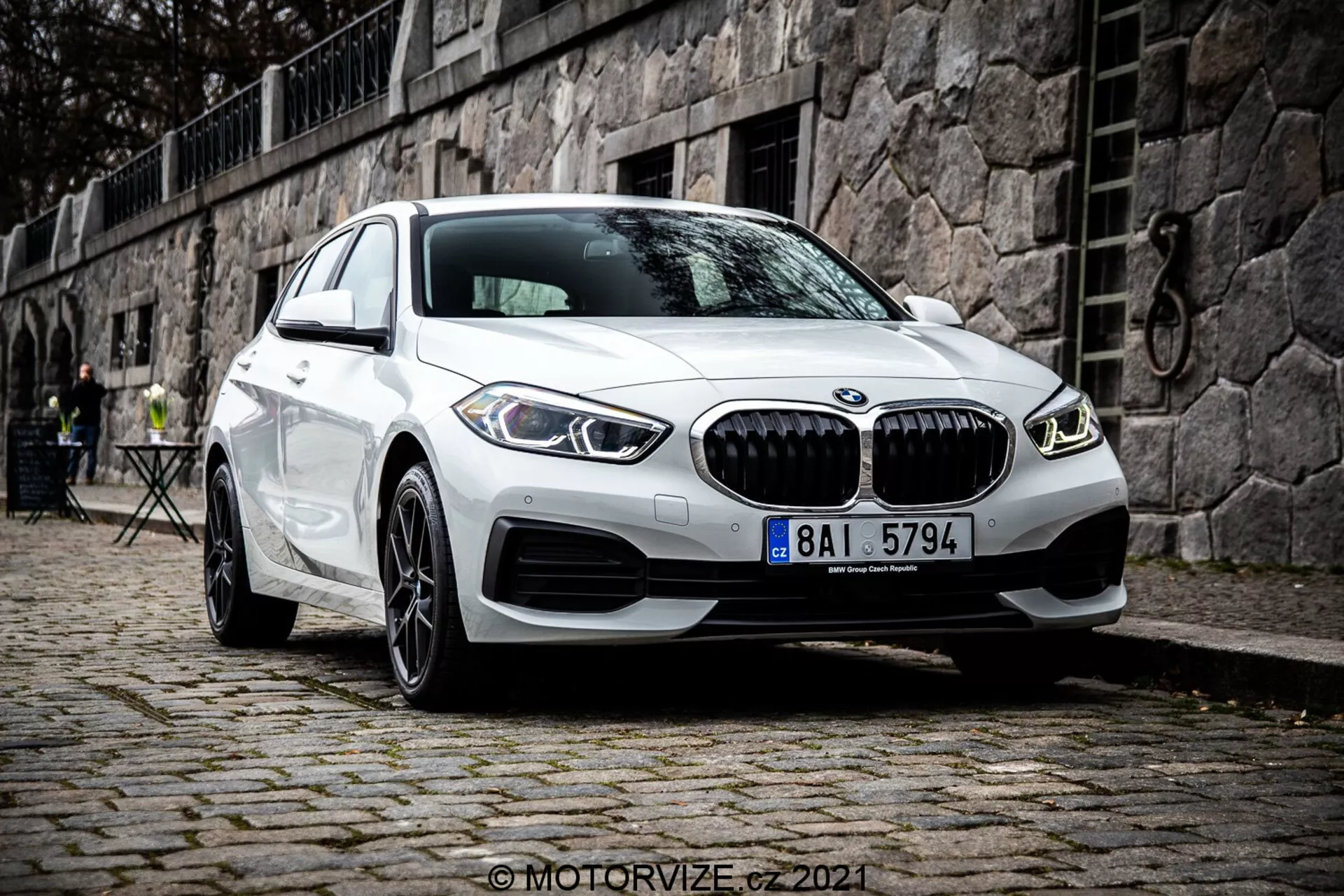 Nowoczesny biały BMW Serii 1 (BMW 116i) model 2019 z charakterystycznym grillem nerkowym, podwójnymi okrągłymi światłami do jazdy dziennej LED i sportowym designem zderzaka przedniego, zaparkowany na brukowanej powierzchni w historycznym europejskim otoczeniu z kamiennym murem i meblami kawiarnianymi na zewnątrz w tle.