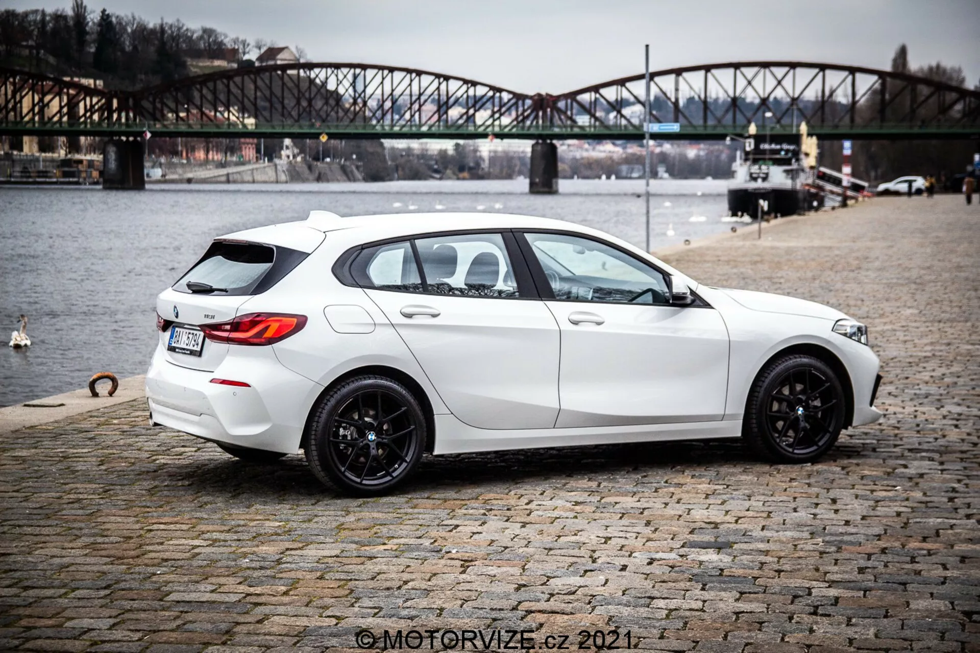 Obraz białego hatchbacka BMW 1er-Reihe 116i z 2019 roku z widokiem na trzy czwarte od tyłu, podkreślający nowoczesny, elegancki design z sportową sylwetką, czarnymi wieloramiennymi felgami, lekko przyciemnianymi szybami i praktycznymi funkcjami, takimi jak odmrażacz tylnej szyby i wycieraczka. Samochód prezentuje ostre linie i kontury, podwójny układ wydechowy, subtelny tylny spojler dachowy, kamerę cofania i czujniki parkowania. Zaparkowany na brukowanej powierzchni w pobliżu zbiornika wodnego z miejskim mostem w tle, sugeruje kontekst życia na świeżym powietrzu.