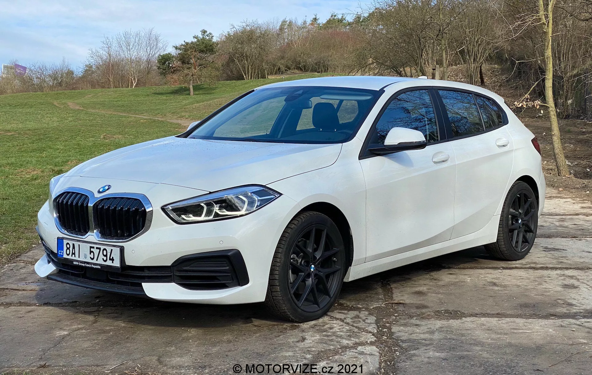 Obraz przedstawia nowoczesny biały hatchback BMW Serii 1 (BMW 116i) umieszczony na zewnątrz z naturalnym, trawiastym tłem. Uchwycony z przedniego widoku trzy-czwarte, faworyzując stronę kierowcy, ukazuje charakterystyczną nerkową kratkę pojazdu, eleganckie kanciaste reflektory LED, czarne felgi aluminiowe, dobrze zdefiniowaną fałdę boczną oraz zintegrowane kierunkowskazy na lusterkach bocznych. Przedni zderzak wyposażony jest w czujniki parkowania, podczas gdy można wnioskować o panoramicznym dachu szklanym i możliwej przedniej kamerze. Samochód jest zaparkowany na brukowanej powierzchni, zajmując większość kadru, co sugeruje nowoczesny design motoryzacyjny i zaawansowane funkcje technologiczne.