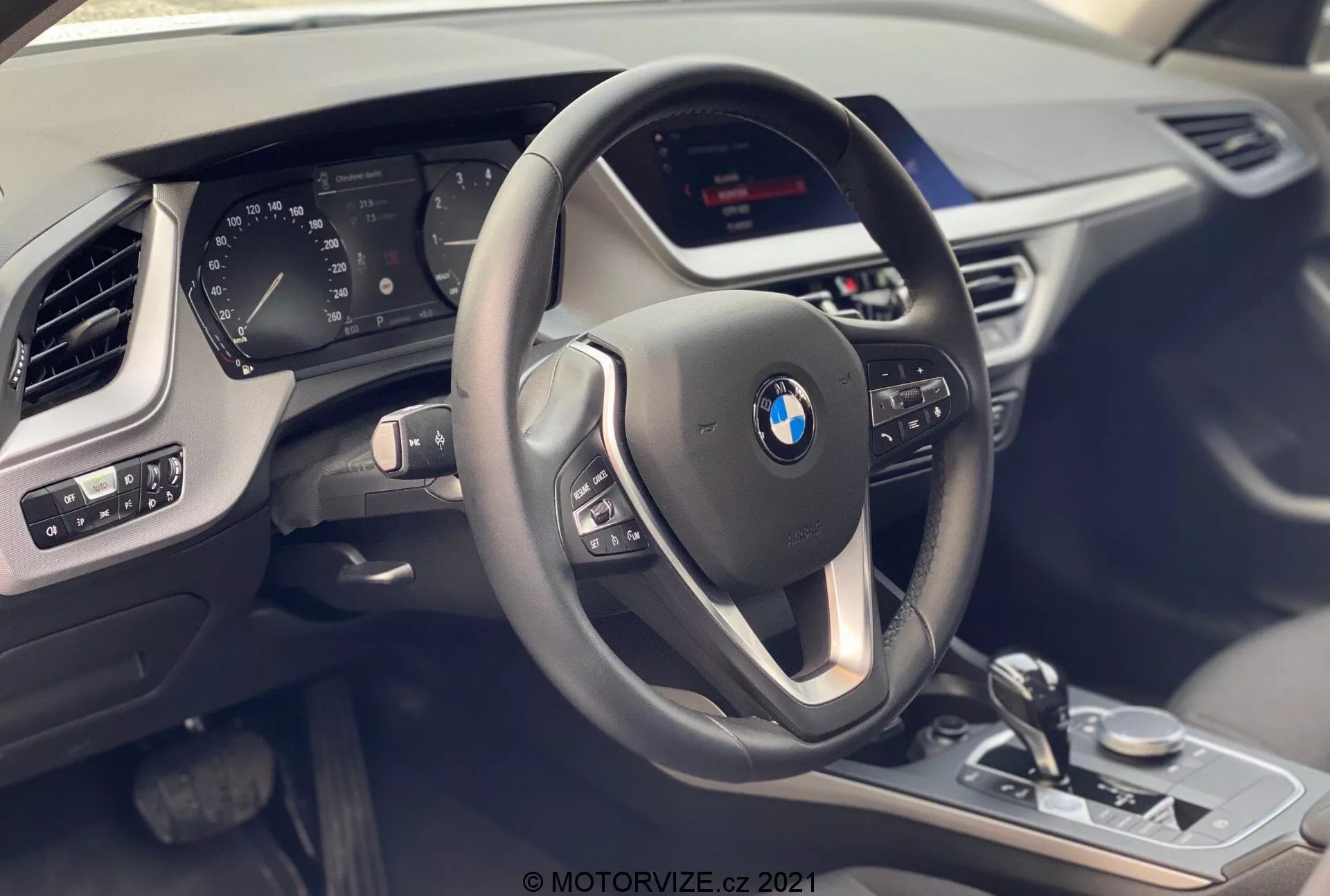 Widok wnętrza BMW Serii 1 2019 (BMW 116i) z siedzenia pasażera, podkreślający kokpit kierowcy z nowoczesną, trójramienną kierownicą wyposażoną w wielofunkcyjne przyciski oraz skórzane pokrycie z kontrastującymi przeszyciami. Za kierownicą widoczny jest częściowo cyfrowy zestaw wskaźników pokazujący różne parametry pojazdu. Konsola centralna mieści niewidoczny ekran infotainmentu oraz widoczne elementy sterujące, takie jak selektor biegów, pokrętło sterowania iDrive, selektor trybu jazdy oraz kontrolki czujników parkowania, wszystkie wykończone metalicznymi akcentami i błyszczącymi czarnymi panelami. Panel drzwi kierowcy pokazuje regulacje lusterka i sterowanie szybami. Widoczna jest także część nawiewu po stronie pasażera oraz tekstura deski rozdzielczej, podkreślająca luksus, technologię oraz nowoczesny design.