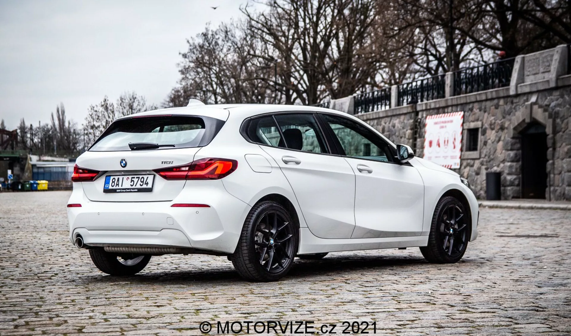 Wysokiej jakości zdjęcie białego hatchbacka BMW Serii 1 2019 128ti zrobione pod kątem trzech czwartych od tyłu, podkreślające prawą stronę. Samochód zaparkowany jest na brukowanym chodniku, z kamiennym murem w tle, co podkreśla jego sportowy wygląd z tylnym spojlerem, podwójnym układem wydechowym, tylnym dyfuzorem, tylne światła LED, przyciemnianymi szybami i ciemnymi alufelgami. Rozproszone światło, brak ostrych cieni oraz wyraźne detale sugerują pochmurne otoczenie, co daje autentyczne odwzorowanie koloru i cech pojazdu. Na obrazku widoczny jest znak wodny 'MotorVize.cz 2021'.