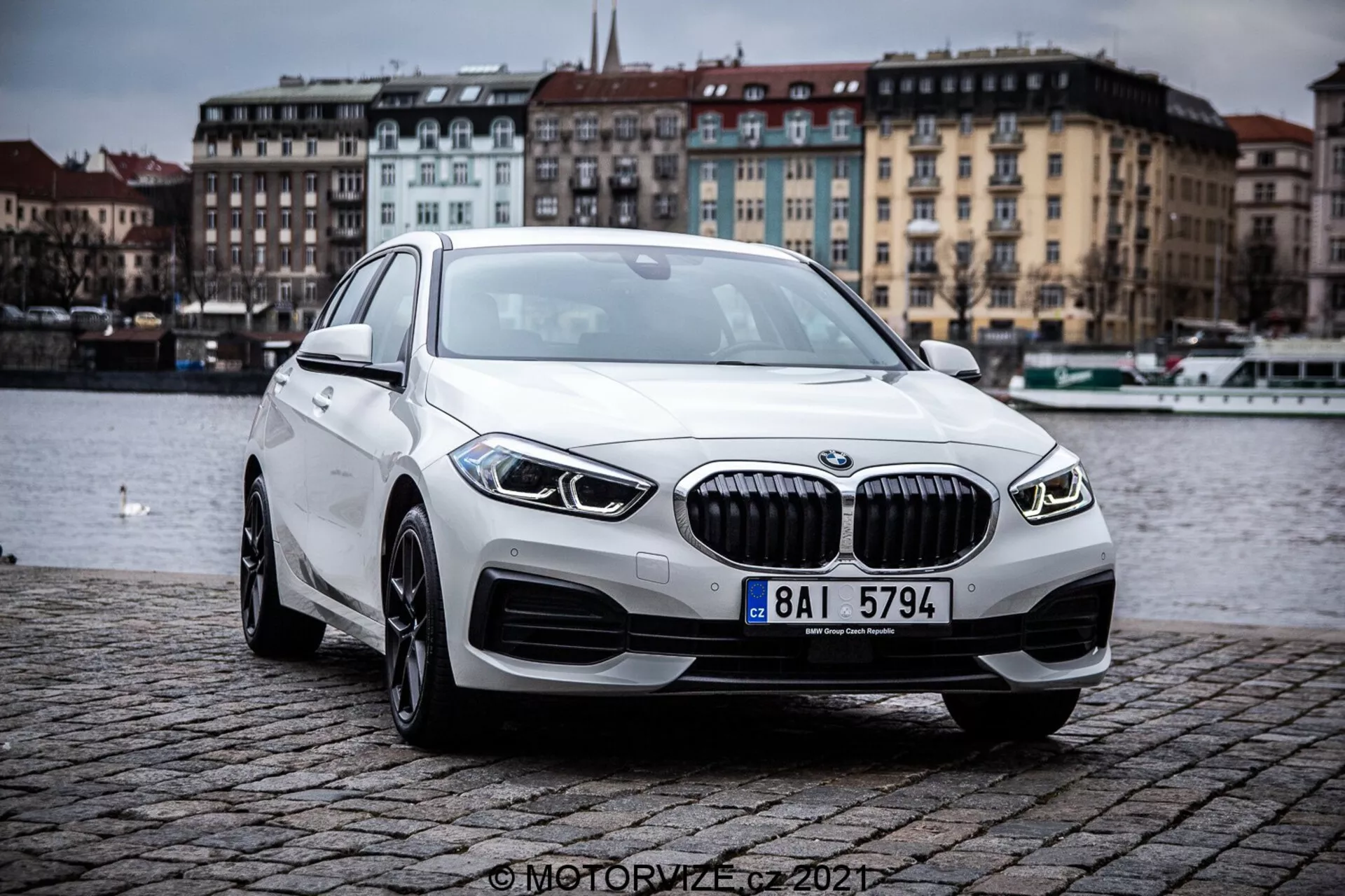 Obraz przedstawiający BMW 1er-Reihe 2019 (BMW 116i) z perspektywy trzy czwarte z przodu, podkreślający kontury i elementy designu nowoczesnego luksusowego sedana. Samochód znajduje się na zewnątrz na brukowanym nasypie w pobliżu zbiornika wodnego, pod pochmurnym niebem. Wyraźne cechy obejmują kratkę z pionowymi żaluzjami, eleganckie reflektory LED, zintegrowane światła przeciwmgielne, aluminiowe koła i aerodynamiczny profil.