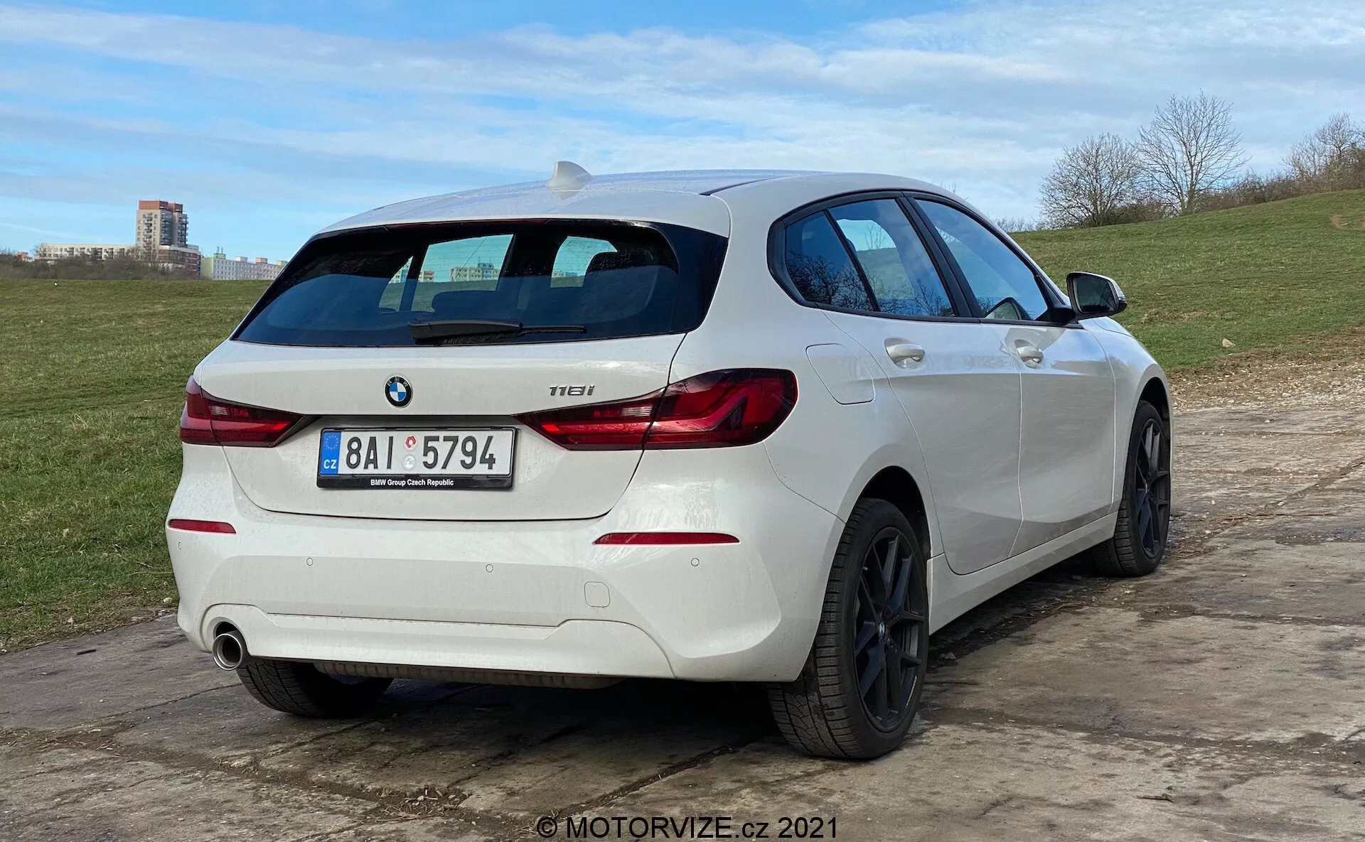 Widok tylnej części z trzech czwartych na białego hatchbacka BMW 1 Series 118i, zaparkowanego na nierównym terenie. Samochód ma charakterystyczne tylne światła w kształcie litery 'L', emblemat BMW oraz plakietkę modelu z napisem '118i'. Posiada podwójny układ wydechowy z jedną widoczną rurą po prawej stronie tylnego zderzaka. Pojazd wyposażony jest w wieloszprychowe czarne felgi aluminiowe i tylnią wycieraczkę. Spoiler dachowy dodaje sportowego wyglądu, a profil boczny ujawnia lekko przyciemniane szyby, fałdy nadwozia oraz pasujące do kolorystyki samochodu klamki i lusterka. Niski kąt kamery podkreśla postawę samochodu i design kół.