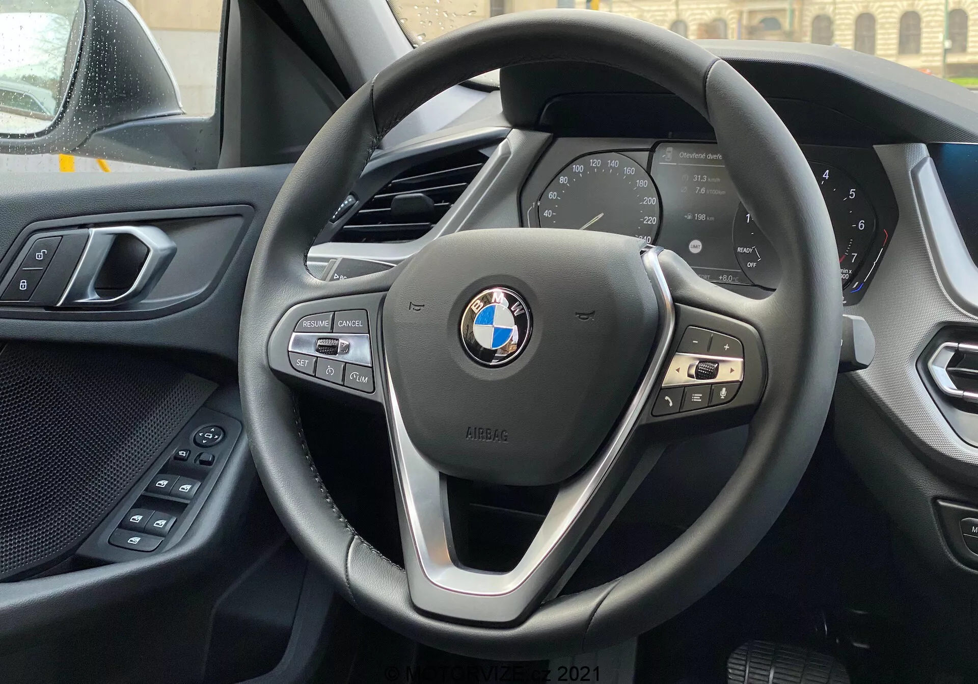 Wnętrze BMW serii 1 z 2019 roku (BMW 116i) widok z fotela kierowcy koncentruje się na nowoczesnej, trójramiennej kierownicy BMW z przyciskami do obsługi multimediów, telefonu i tempomatu oraz cyfrowym zestawie wskaźników pokazującym podstawowe parametry pojazdu, takie jak prędkość i obroty. Górna krawędź konsoli centralnej jest widoczna w tle, po lewej widać część drzwi kierowcy z elektrycznymi przyciskami komfortu. Deska rozdzielcza i panel drzwiowy prezentują połączenie miękkich materiałów i wysokiej jakości wykonania. Emblemat BMW i znaczek iPERFORMANCE są dobrze widoczne na kierownicy, co sugeruje efektywność lub technologię hybrydową.