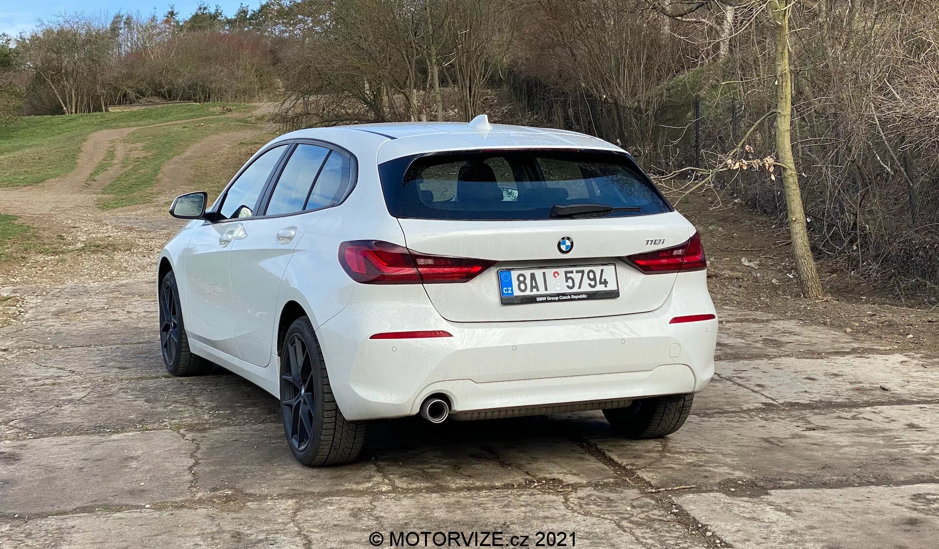 Zdjęcie przedstawia białego hatchbacka BMW 1er-reihe 2019 (BMW 116i) widzianego pod kątem tylnej trzeciej części auta. Tył pojazdu, w tym poziomo zorientowane światła tylne, podwójny układ wydechowy i czujniki parkowania zintegrowane z tylnym zderzakiem, jest wyraźnie widoczny. Na tylnej klapie widoczne są emblemat BMW i oznaczenie modelu. Samochód znajduje się na żwirowej powierzchni z naturalnym, wiejskim tłem. Zdjęcie sugeruje tematy motoryzacyjne takie jak design pojazdów, współczesne modele aut, treści dotyczące BMW, układy wydechowe i technologię wspomagania parkowania. Obecność znaku wodnego MOTORVIZE.cz 2021 wskazuje na pochodzenie z recenzji lub publikacji motoryzacyjnej.
