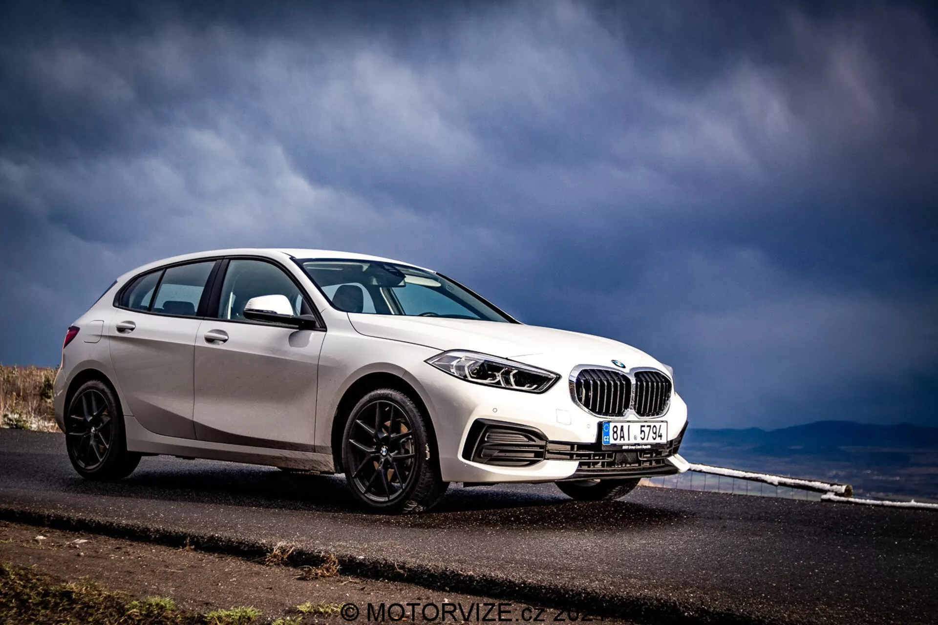 BMW 1 Series 2019 (BMW-116i) uchwycony w dramatycznym przednim widoku trzech czwartych, ukazujący jego przedni i boczny profil. Samochód, wykończony w jasnym białym kolorze z kontrastującymi czarnymi akcentami, takimi jak obudowy lusterek, listwy okienne i wieloramienne koła, znajduje się po lewej stronie kadru na tle asfaltowej drogi prowadzącej w oddali i burzowego nieba. Widoczne elementy obejmują szczegółowe reflektory z okrągłymi elementami, wyraźną nerkę, przedni zderzak z otworami wentylacyjnymi i potencjalnymi czujnikami parkowania.