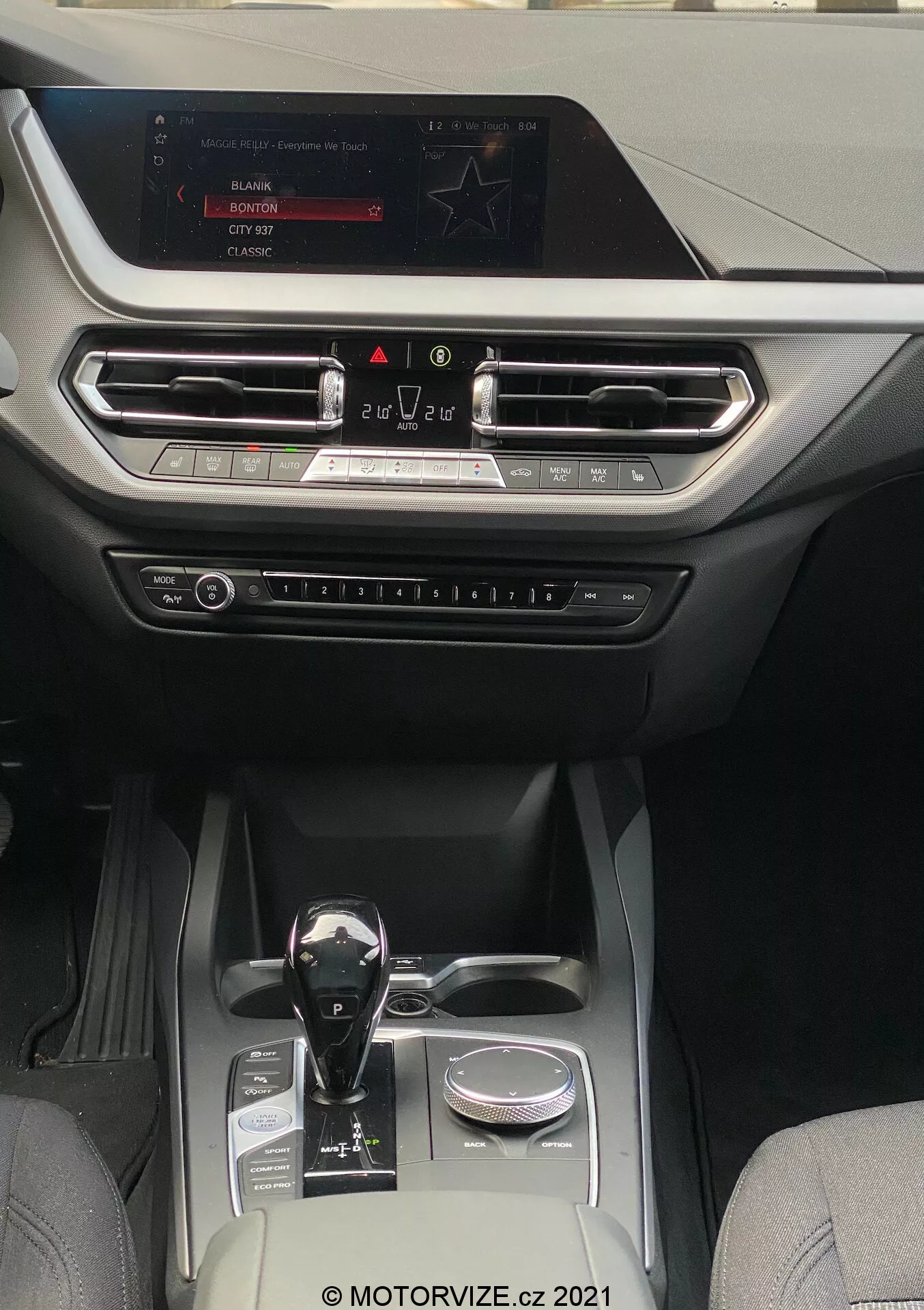 Widok wnętrza BMW 1er-reihe z 2019 roku (BMW-116i), ukazujący nowoczesną i luksusową kabinę skierowaną na konsolę środkową i obszar deski rozdzielczej. Perspektywa jest z tylnych siedzeń, patrząc do przodu na ekran infotainment zamontowany na górze deski rozdzielczej. Wyświetlacz pokazuje opcje radia, mediów i ustawień samochodu. Poniżej ekranu znajdują się poziome otwory wentylacyjne z metalicznymi akcentami i dekoracyjną listwą, które rozciągają się na całą szerokość deski rozdzielczej. Poniżej otworów wentylacyjnych widoczne są fizyczne przyciski i pokrętła do regulacji klimatyzacji oraz innych ustawień. Automatyczna dźwignia zmiany biegów ma błyszczące czarne wykończenie z metalicznymi akcentami, umieszczona obok pokrętła obrotowego. Konsola środkowa zawiera dwa uchwyty na kubki, mały schowek i srebrny element dekoracyjny, co świadczy o dobrze wyposażonej kabinie łączącej skórę i wysokiej jakości tkaniny syntetyczne, odpowiedniej dla pojazdów średniej i wyższej klasy, które koncentrują się na technologii i komforcie.