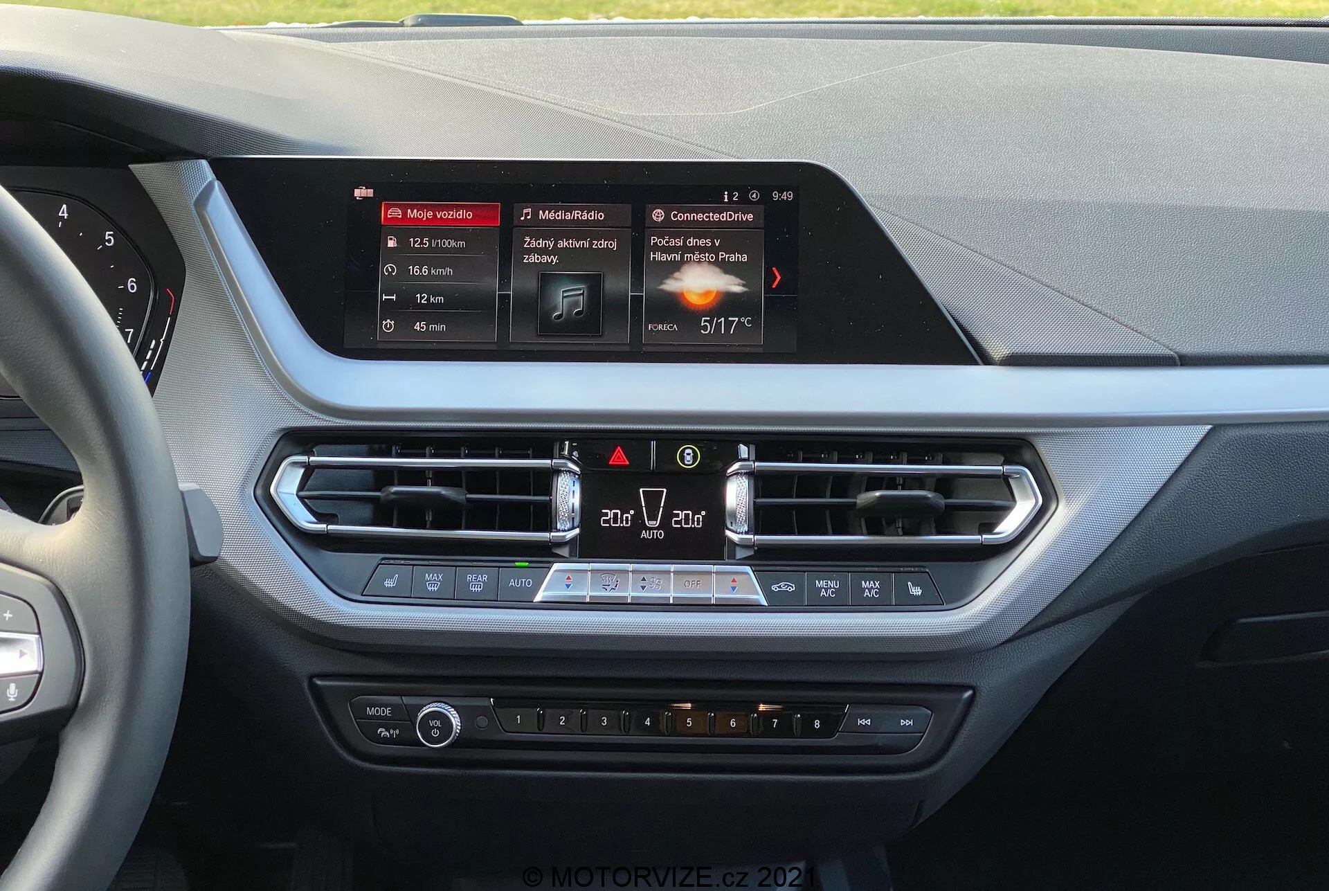 Wnętrze BMW serii 1 2019 (BMW-116i) z perspektywy kierowcy, prezentujące konsolę środkową deski rozdzielczej. Centralnie umieszczony jest podświetlany, prostokątny wyświetlacz dotykowy systemu infotainment, pokazujący nawigację, multimedia i szczegóły klimatyzacji. Poniżej ekranu znajdują się otwory wentylacyjne z metalowymi wykończeniami, kontrolki zagrożenia i recyrkulacji powietrza, a następnie panel sterowania klimatem z pokrętłami temperatury, prędkości wentylatora i podgrzewanymi siedzeniami. Materiały cechują miękkie w dotyku powierzchnie i chromowane akcenty, sugerujące skupienie się na komforcie kierowcy i pasażera z przodu.