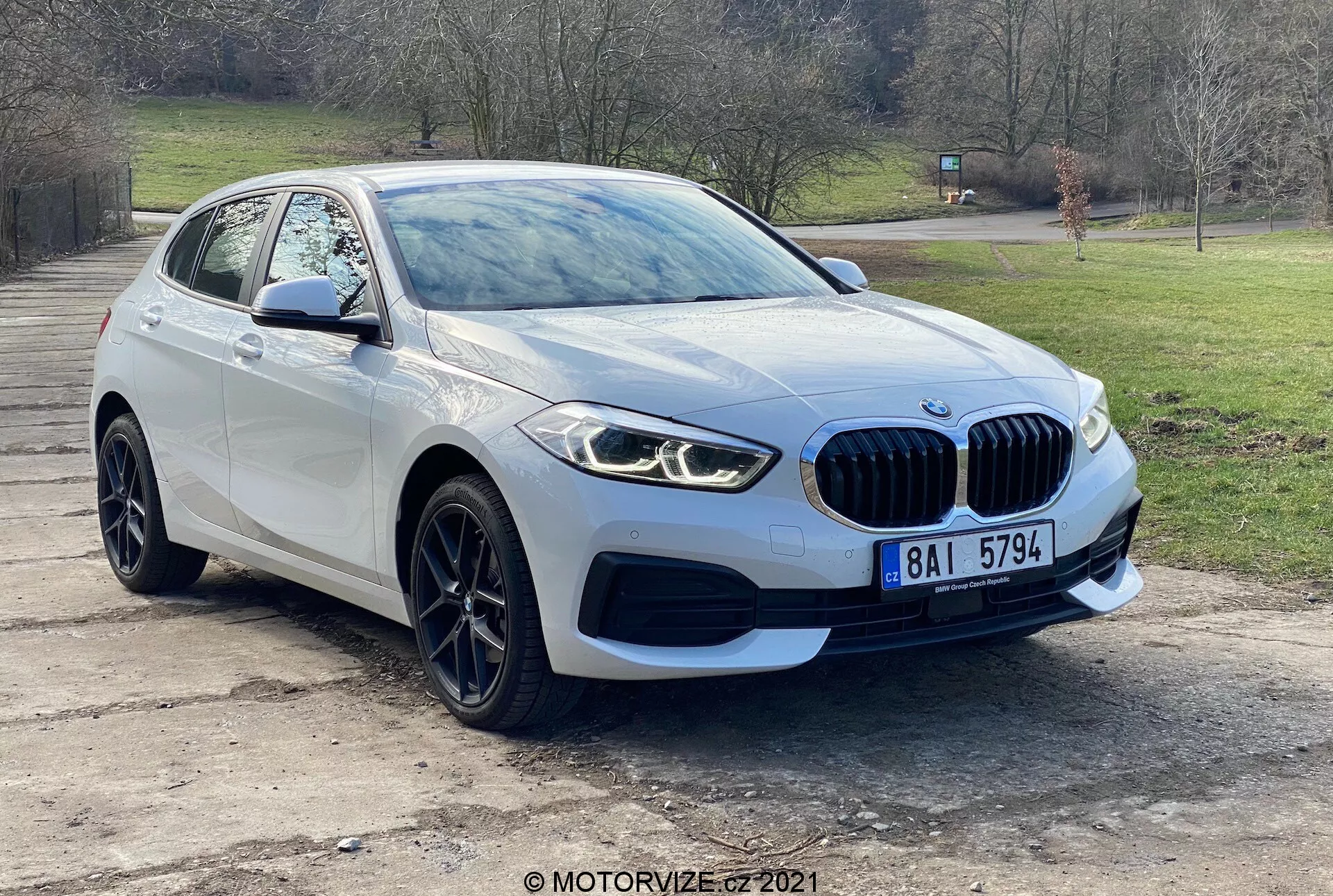 Nowoczesny pojazd BMW 116i z serii 1 z 2019 roku sfotografowany z perspektywy przedniego trzech czwartych, prezentujący przedni grill z emblematem, eleganckie światła LED, światła przeciwmgielne, przedni zderzak i aerodynamiczne linie maski prowadzące do przedniej szyby. Koło i błotnik po stronie kierowcy, z lusterkiem bocznym z wbudowanymi kierunkowskazami, podkreślają zaawansowany design i technologię czujników parkowania pojazdu. Uchwycone na zewnątrz z tłem drzew i trawy, auto ma aluminiowe koła i charakterystyczny grill w kształcie nerki, co wskazuje na luksus i wydajność.