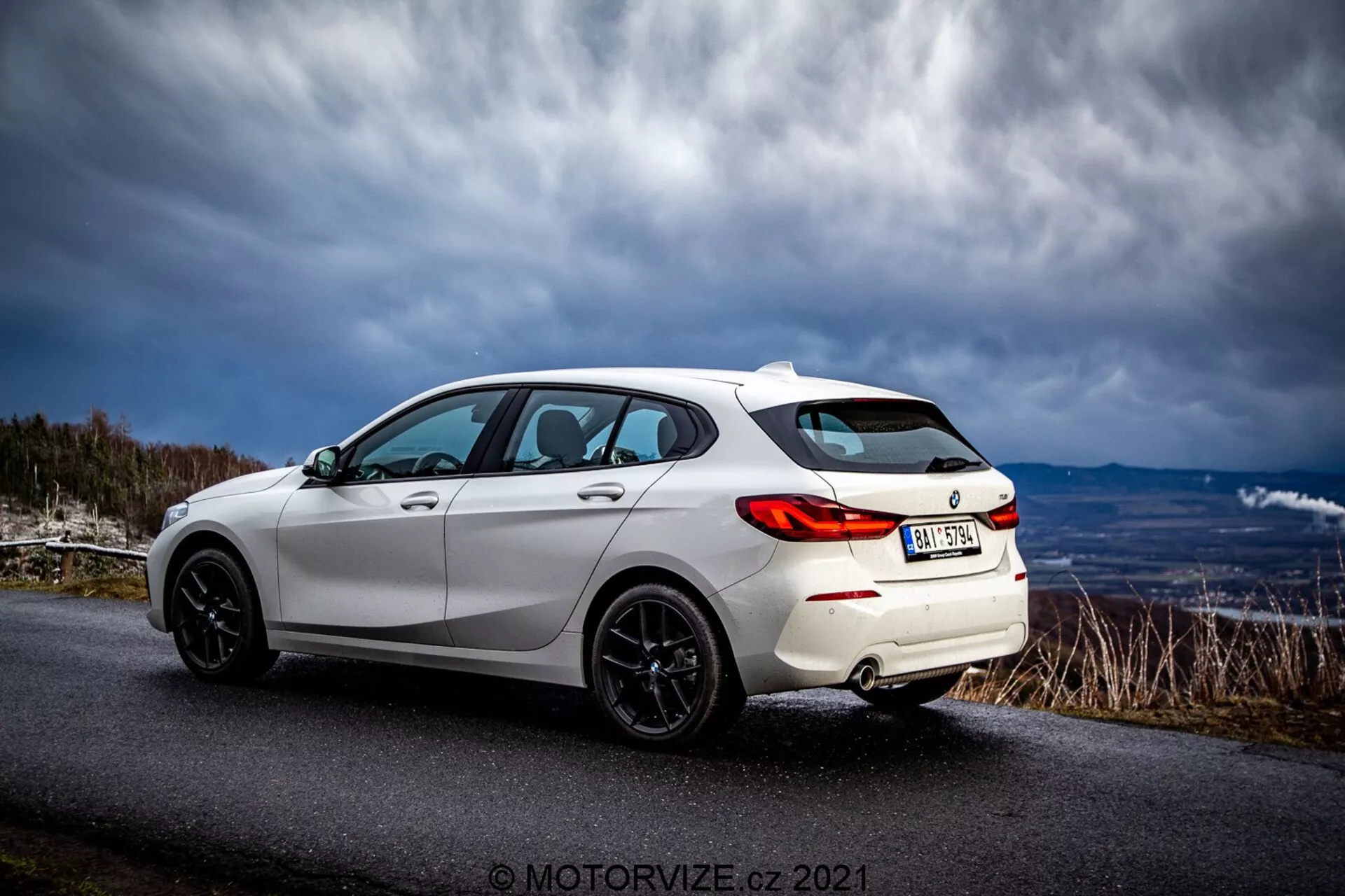 Biały, nowoczesny hatchback BMW 116i 2019 uchwycony w widoku trzech czwartych tyłu. Sportowe cechy zewnętrzne obejmują tylne światła LED, czarne wieloramienne felgi aluminiowe, tylny spojler, przyciemniane szyby i stylowe linie nadwozia podkreślające jego aerodynamiczny design. Pojazd jest zaparkowany na asfaltowej drodze w wiejskiej lub górskiej okolicy pod dramatycznym niebem.