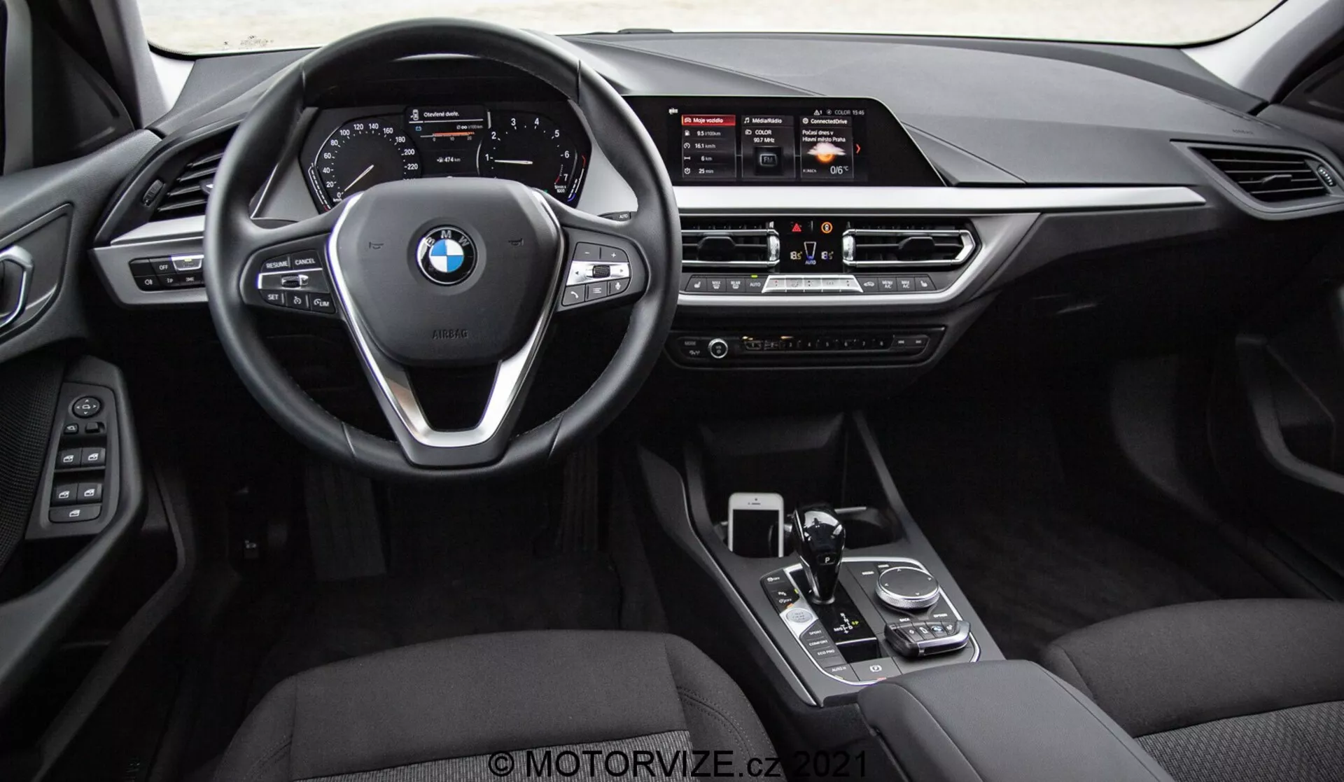 Widok wnętrza BMW Serii 1 2019 (BMW 116i) uchwycony z tylnych siedzeń, ukazujący cały przedni kokpit, kierownicę z logo BMW i przyciskami kontrolnymi oraz konsolę środkową. Widoczne elementy to szeroki wyświetlacz infotainment, ręczne sterowanie klimatyzacją, nawiewy powietrza, dźwignię zmiany biegów, pokrętło i touchpad. Wnętrze wykończone jest czarną skórą lub skórą ekologiczną z metalowymi i błyszczącymi elementami wykończeniowymi, podkreślającymi luksus i zaawansowaną technologię interfejsu kierowcy.