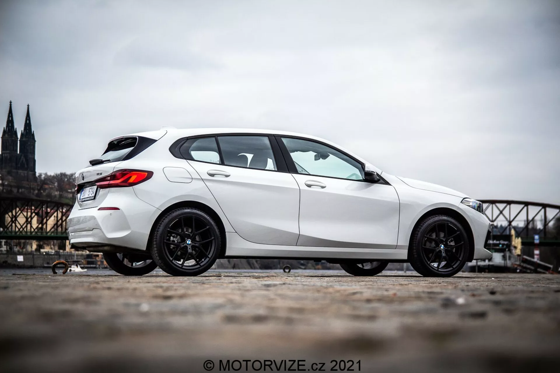 Biały hatchback BMW Serii 1 (BMW 116i) z 2019 roku sfotografowany pod kątem bocznym, pokazujący bok pojazdu od przedniego koła aż do tyłu. Samochód jest ustawiony po lewej stronie obrazu z rozmytym historycznym motywem miejskim, który obejmuje katedrę i most. Zdjęcie podkreśla sportowe, ciemne felgi samochodu i jego dominującą sylwetkę dzięki perspektywie z niższego poziomu i lekkiemu nachyleniu kamery ku górze. Zdjęcie jest wykonane na zewnątrz w ciągu dnia, ukazując szczegóły estetyczne samochodu w naturalnym świetle dziennym.