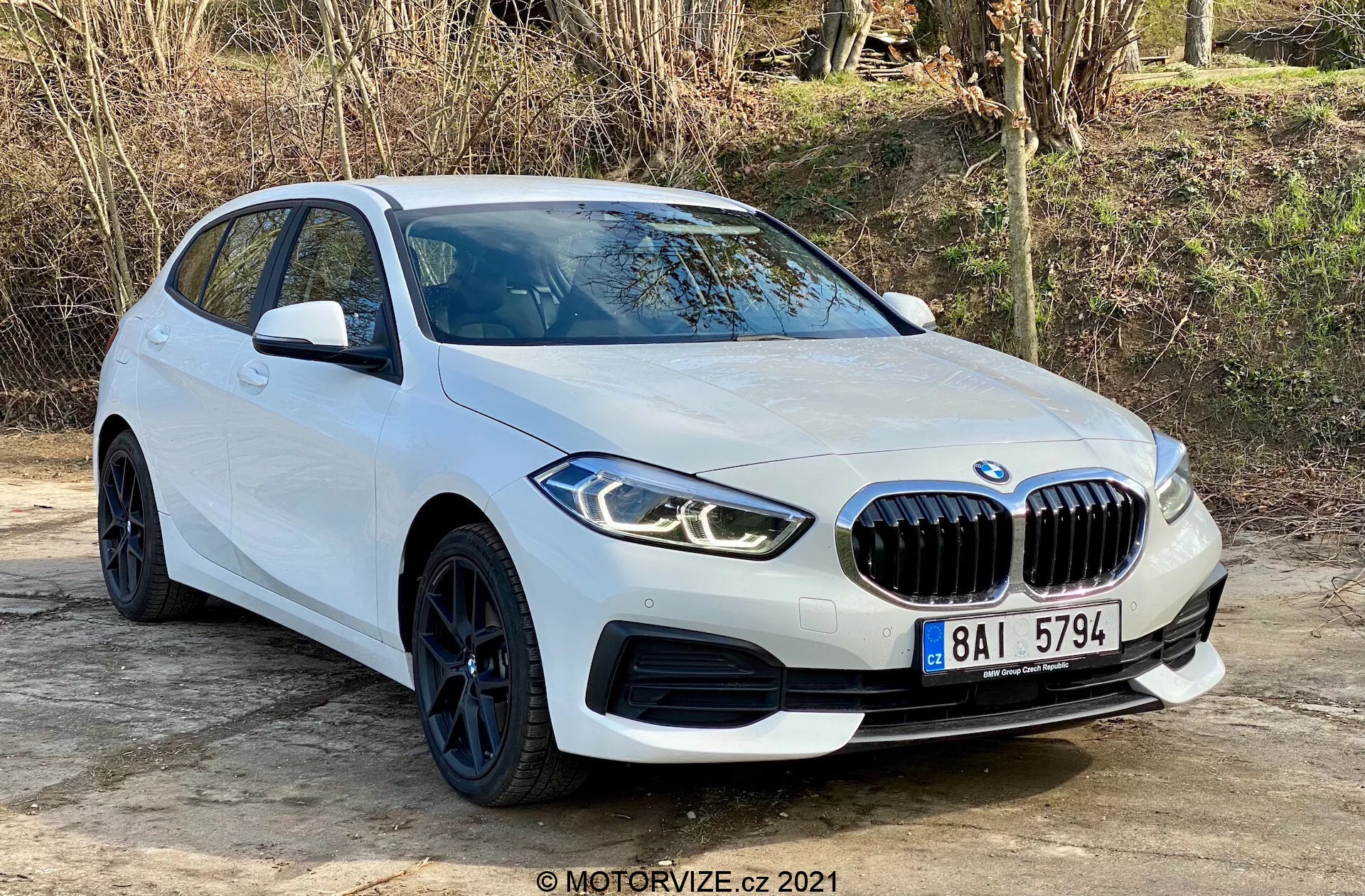 BMW 116i Seria 1 z 2019 roku w białym kolorze, uchwycony pod kątem trzech czwartych, prezentujący jego charakterystyczną nerkową kratkę przednią, kątowe reflektory LED, eleganckie czarne koła z oponami o niskim profilu i aerodynamiczne linie nadwozia. Ustawiony na zewnątrz na płaskiej powierzchni z naturalną roślinnością w tle, ujęcie podkreśla nowoczesny design i sportowe cechy samochodu, w tym czujniki parkowania i lusterka boczne z wbudowanymi kierunkowskazami.