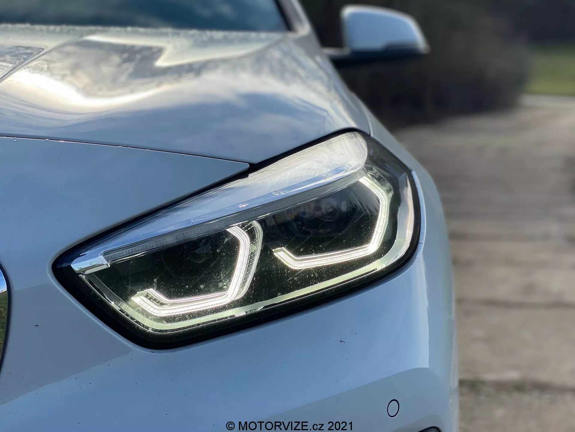 Zbliżenie BMW Serii 1 2019 (BMW 116i) z przedniego ćwierć-kąta, podkreślając lewą stronę i skupiając się na eleganckim, nowoczesnym designie lewego przedniego reflektora z dziennymi światłami LED i przezroczystą pokrywą soczewki. Aerodynamiczne linie maski i błotnika są częściowo widoczne, ukazując jasny metaliczny kolor i błyszczące wykończenie samochodu. Rozmyte tło podkreśla zaawansowane cechy technologiczne reflektora, sugerując lampy LED lub HID, prawdopodobnie wyposażone w adaptacyjną technologię oświetleniową.