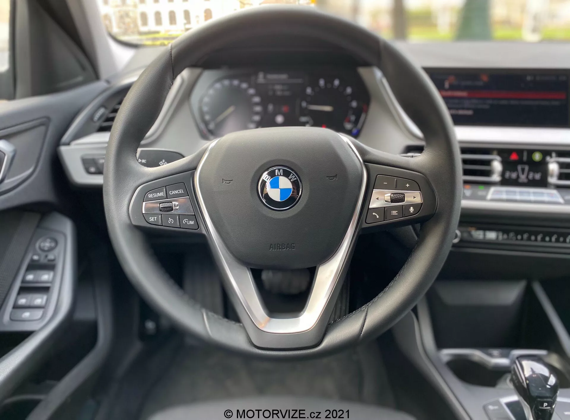 Perspektywa z fotela kierowcy BMW serii 1 2019 (BMW-116i), skupiając się na współczesnej, trójramiennej kierownicy z metalicznym akcentem na dolnym ramieniu i wyraźnym emblematem BMW w centrum. Lewa strona kierownicy zawiera przyciski do obsługi mediów i komunikacji, podczas gdy prawa strona ma przyciski tempomatu i widoczną łopatkę zmiany biegów. W tle widać częściowo widoczny zestaw wskaźników z analogowymi zegarami i ekran infotainment w konsoli środkowej, sugerujący zaawansowane funkcje, takie jak nawigacja GPS i klimatyzacja.