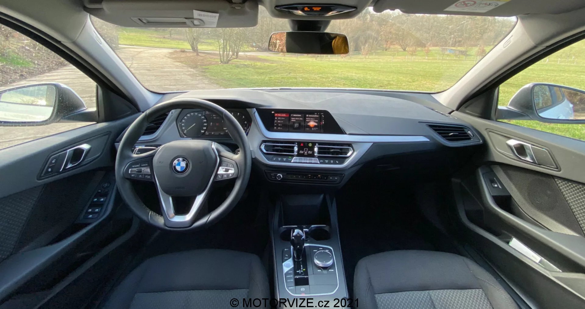 Widok wnętrza z tylnego siedzenia BMW Serii 1 2019 (BMW-116i), ukazując nowoczesną kabinę z wielofunkcyjną, trójramienną kierownicą z logo BMW, wolnostojącym ekranem infotainment, dwustrefową klimatyzacją i automatyczną skrzynią biegów. Użycie skóry, miękkich plastików i metalowych akcentów podkreśla wysoką jakość wykończenia.